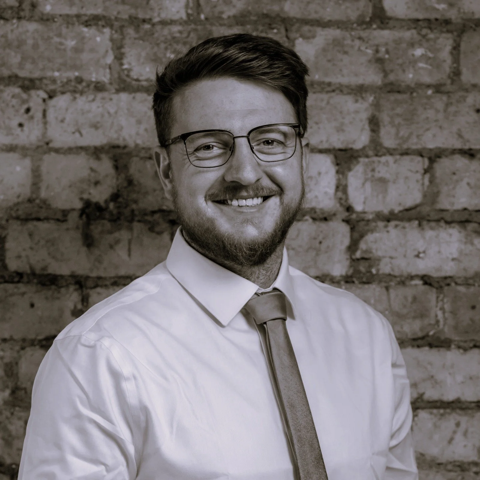A man with glasses, a beard, and a mustache wearing a white dress shirt and tie, smiling in front of a brick wall.