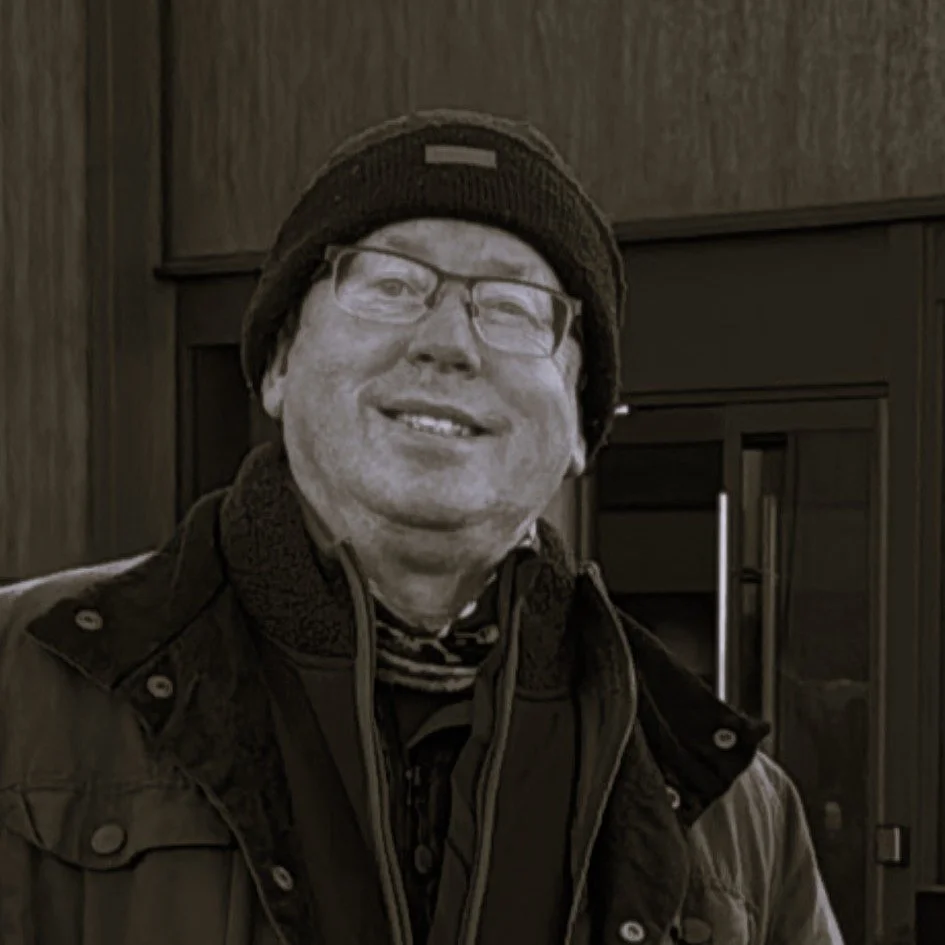 A man wearing glasses, a black beanie, and a layered winter jacket, smiling indoors with wood-paneled walls and a glass door in the background.