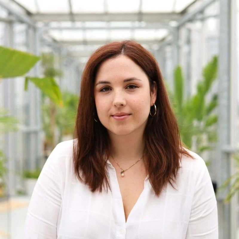 A woman with brown hair, wearing a white shirt and earrings, in a greenhouse setting with green plants in the background.