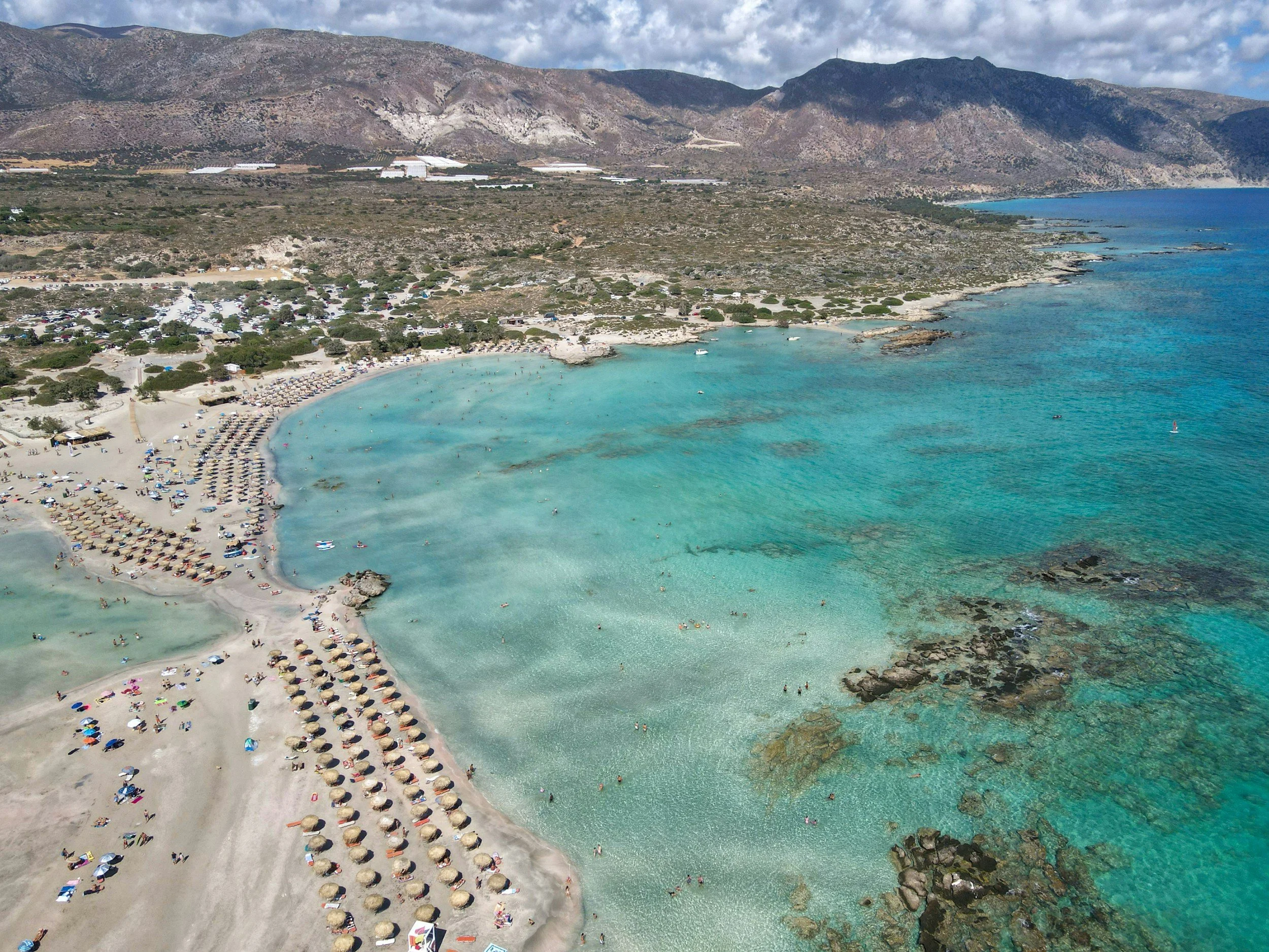 Aerial view of a beach with turquoise water, sandy shore with umbrellas and sunbathers, and mountains in the background.