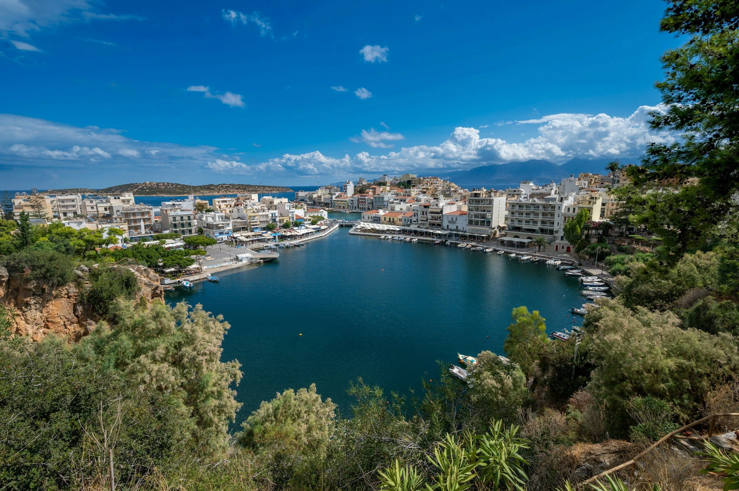 A harbor surrounded by white buildings with boats docked, set against a backdrop of mountains and a partly cloudy sky.