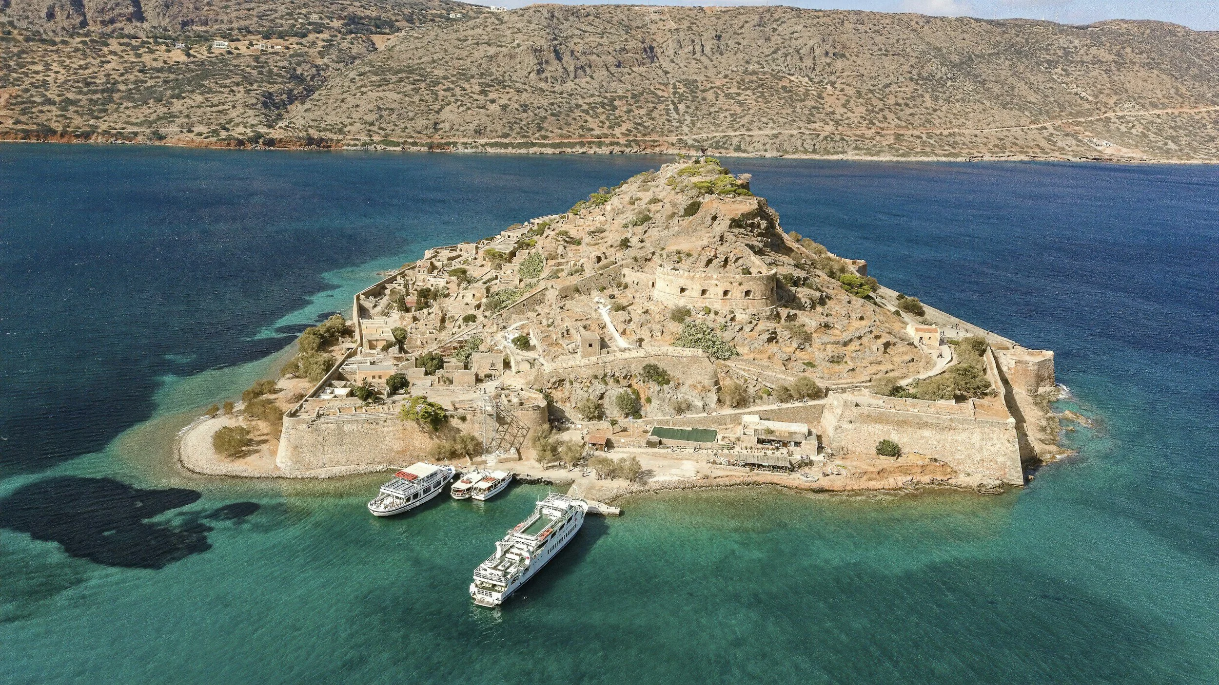 Aerial view of a historic fortress on an island surrounded by turquoise water, with boats docked near the shore, hills in the background.
