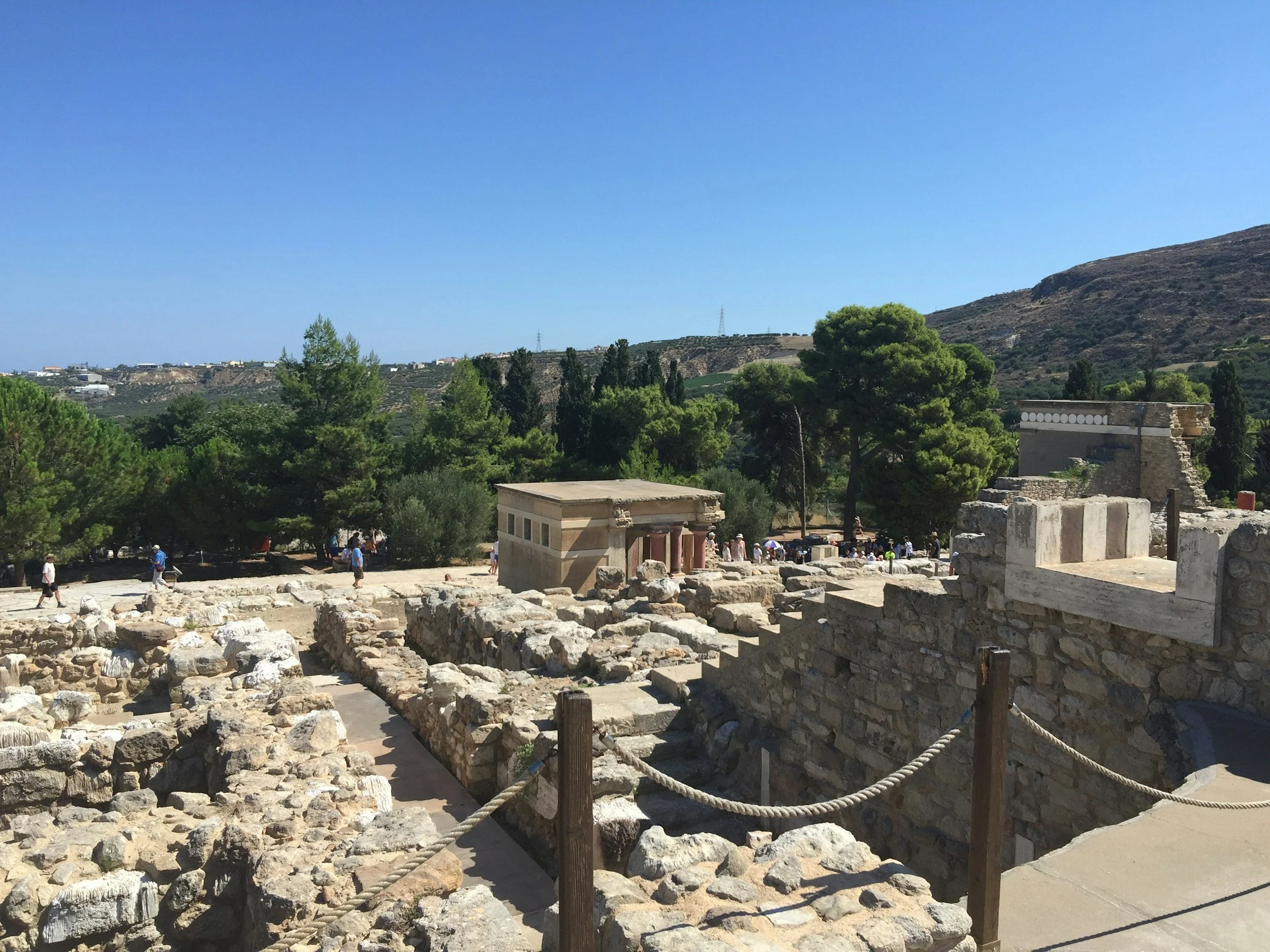 Ancient stone ruins with scattered large stones and small structures, surrounded by green trees under a clear blue sky, with people exploring the archaeological site.