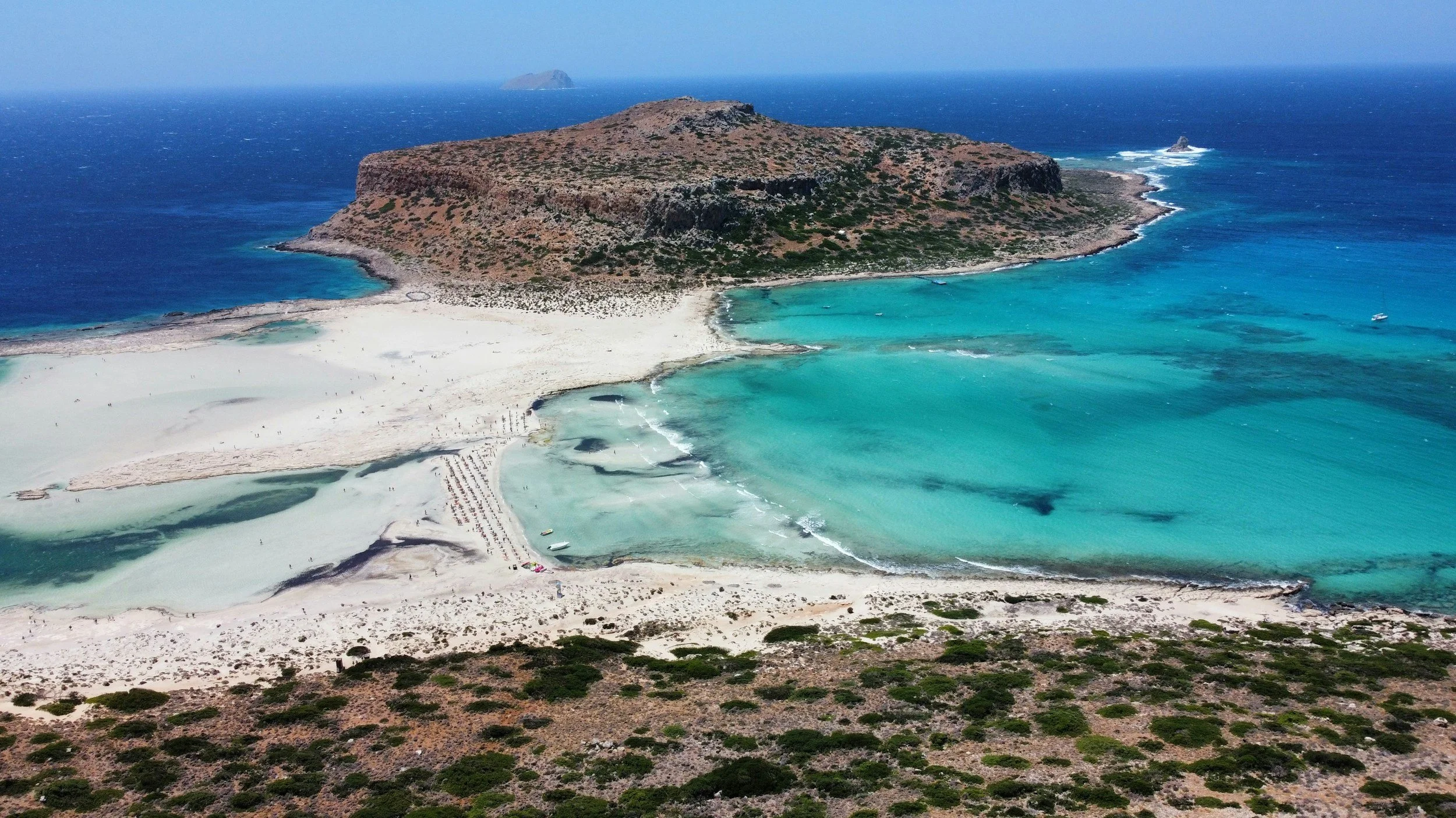 Aerial view of Mykonos, Greece, showing a large sandy beach with turquoise waters, a rocky island in the background, and arid hills with sparse vegetation in the foreground.