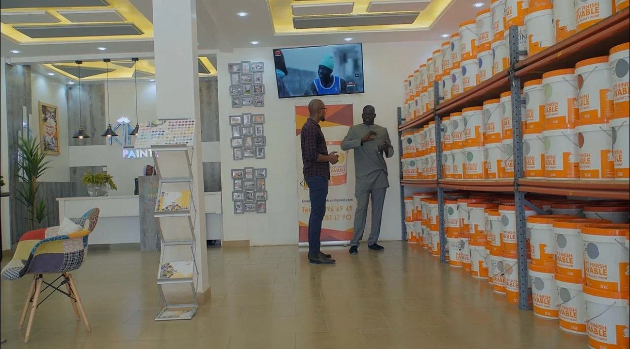 Une salle d'exposition avec deux hommes discutant devant un mur de seaux de peinture orange et blanc. Il y a une télévision au mur, un présentoir à brochures, et un coin salon avec des chaises colorées.