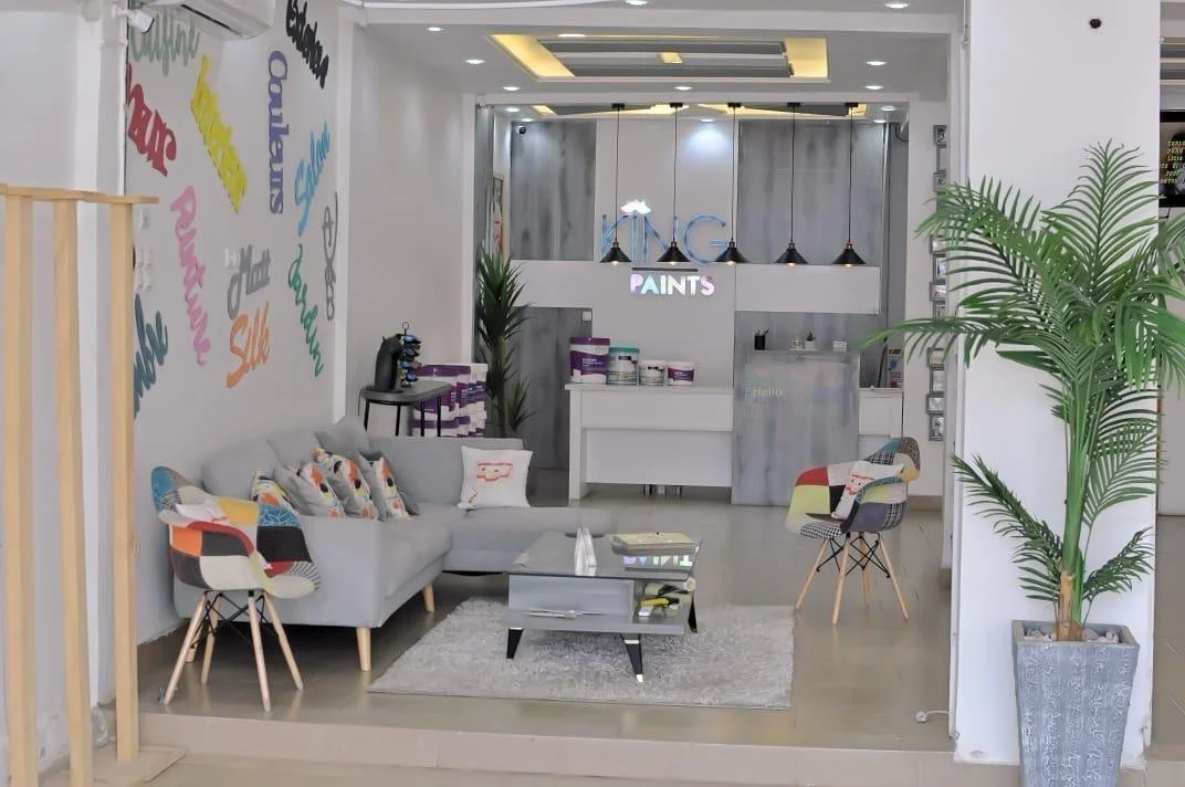 Intérieur d'un salon moderne avec un canapé gris, des fauteuils colorés, un tapis gris, et une grande plante verte. En arrière-plan, un comptoir de peinture avec une enseigne lumineuse "PAINTS".