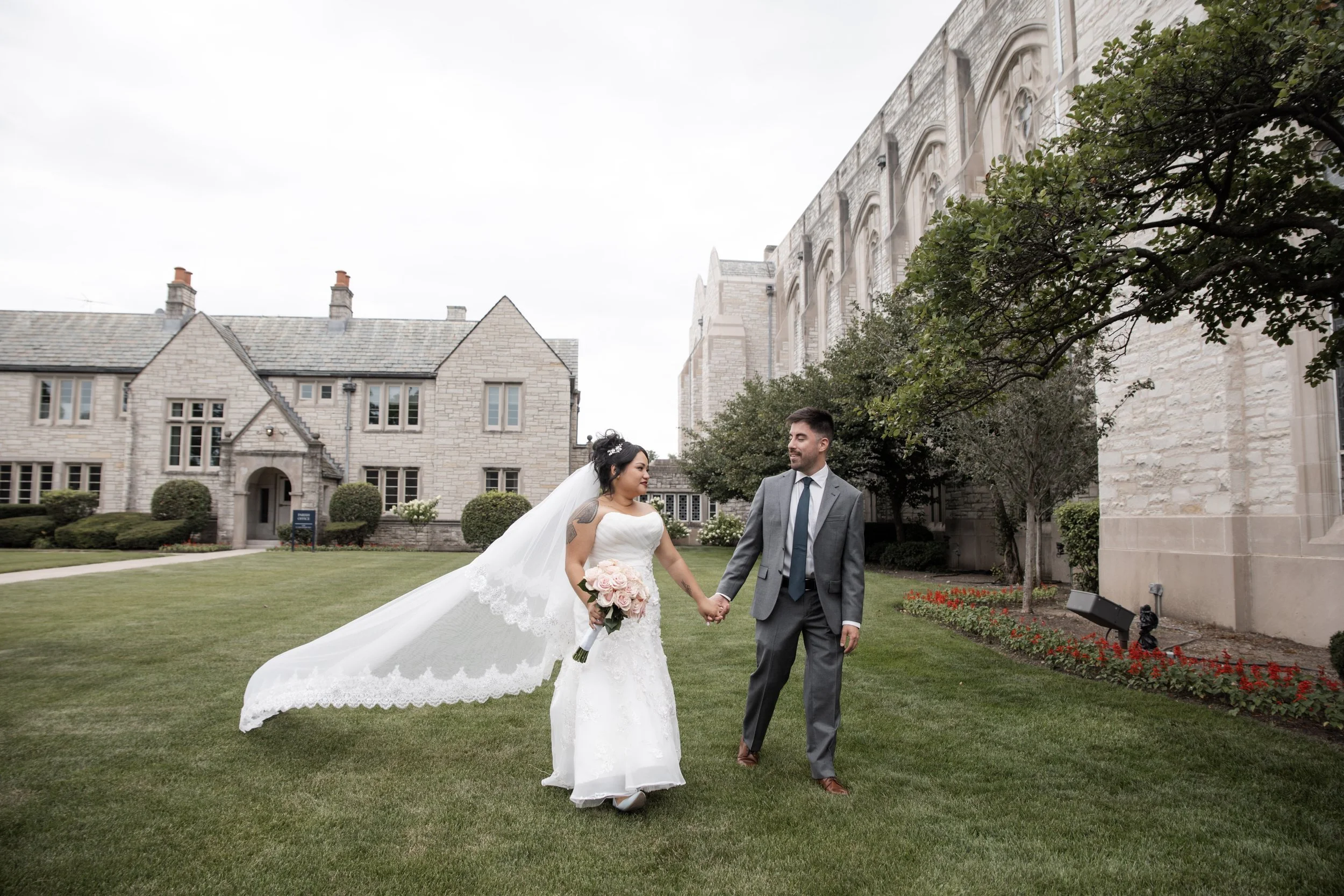A bride and groom holding hands and walking on a green lawn outside a stone building with large windows and trees.
