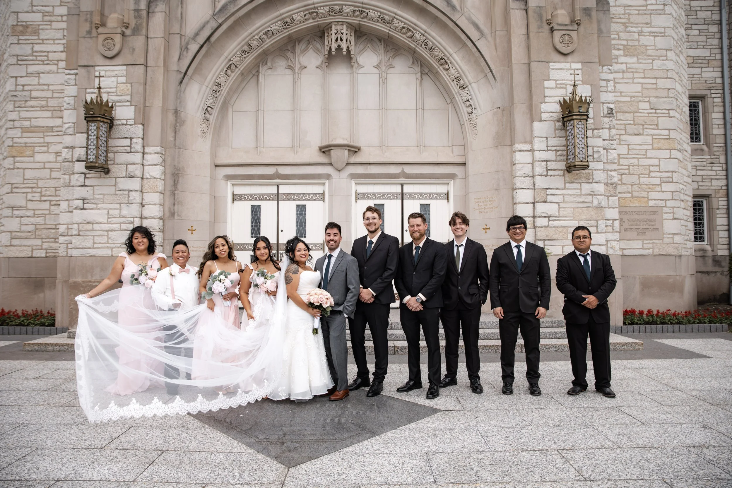 A group of diverse brides and grooms dressed in wedding attire standing in front of a stone church with ornate doors, smiling and posing for a photo.