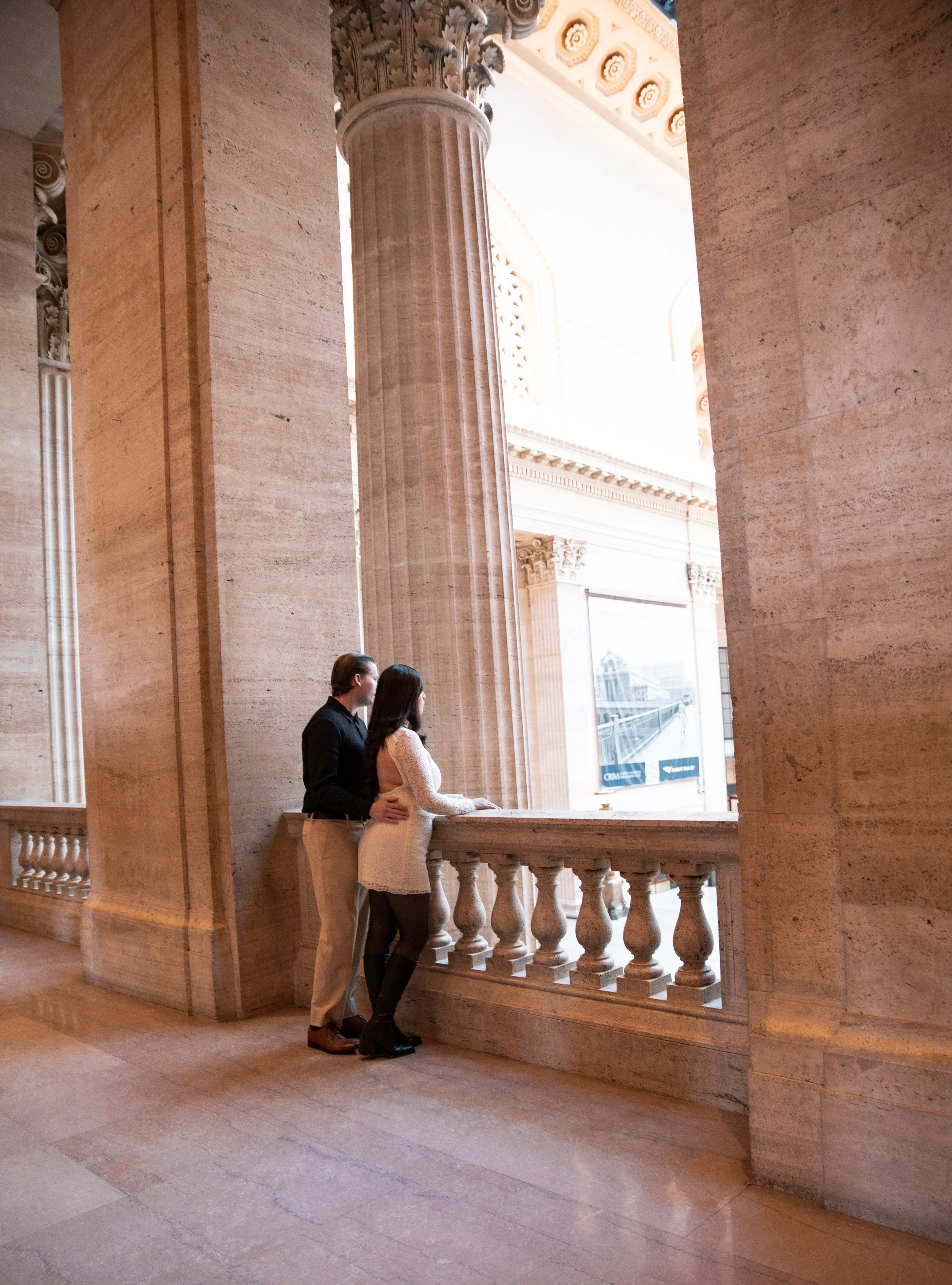 A couple looking out a large window of a grand, historic building with tall columns and intricate architectural details.