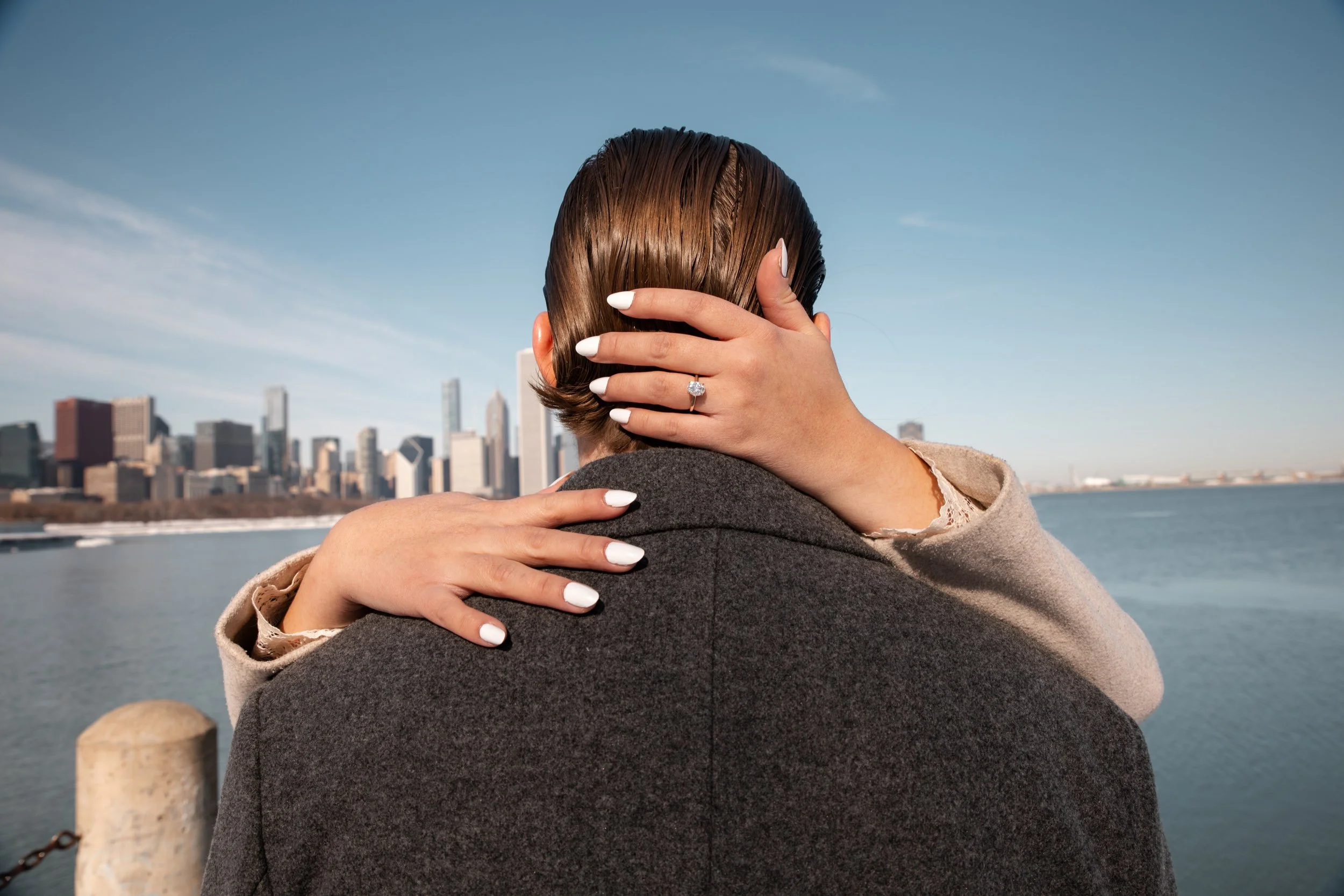 A woman with short, brown hair and a diamond ring on her finger holds a man's head, both embracing in front of a city skyline and water on a clear day.