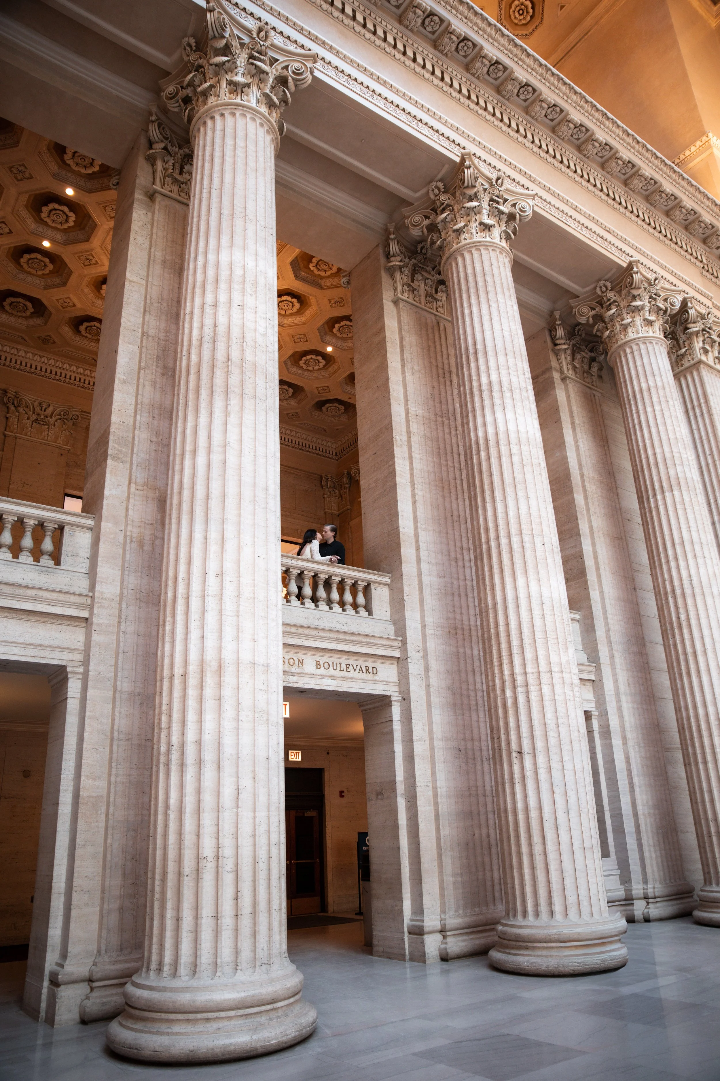 Interior of a grand building with large marble columns, intricate crown molding, and an ornate ceiling with gold accents. Two people are visible on a balcony, possibly engaged in conversation.