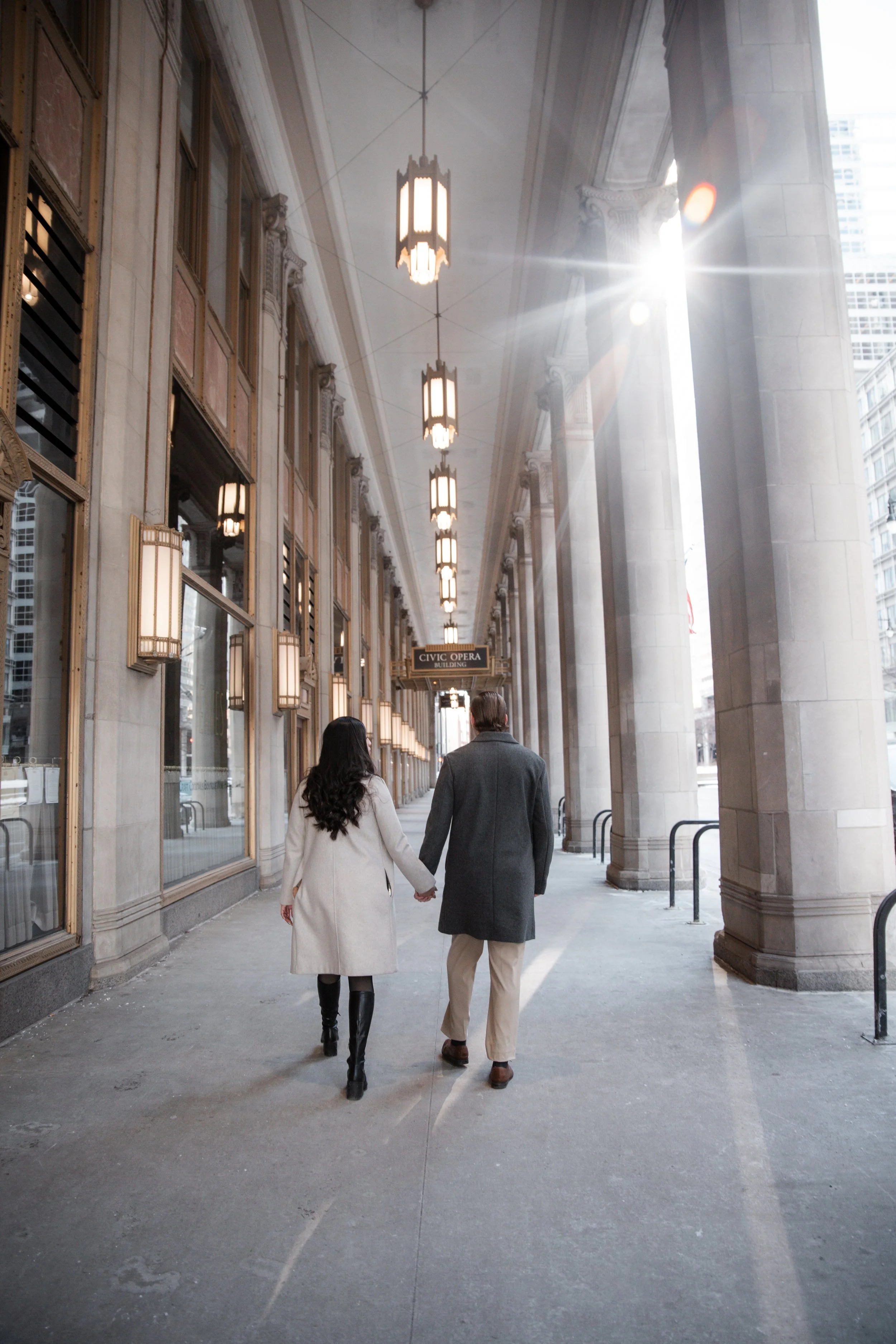 A couple holding hands walking outside a building labeled Civic Opera in Chicago, with sunlight near the top right corner.