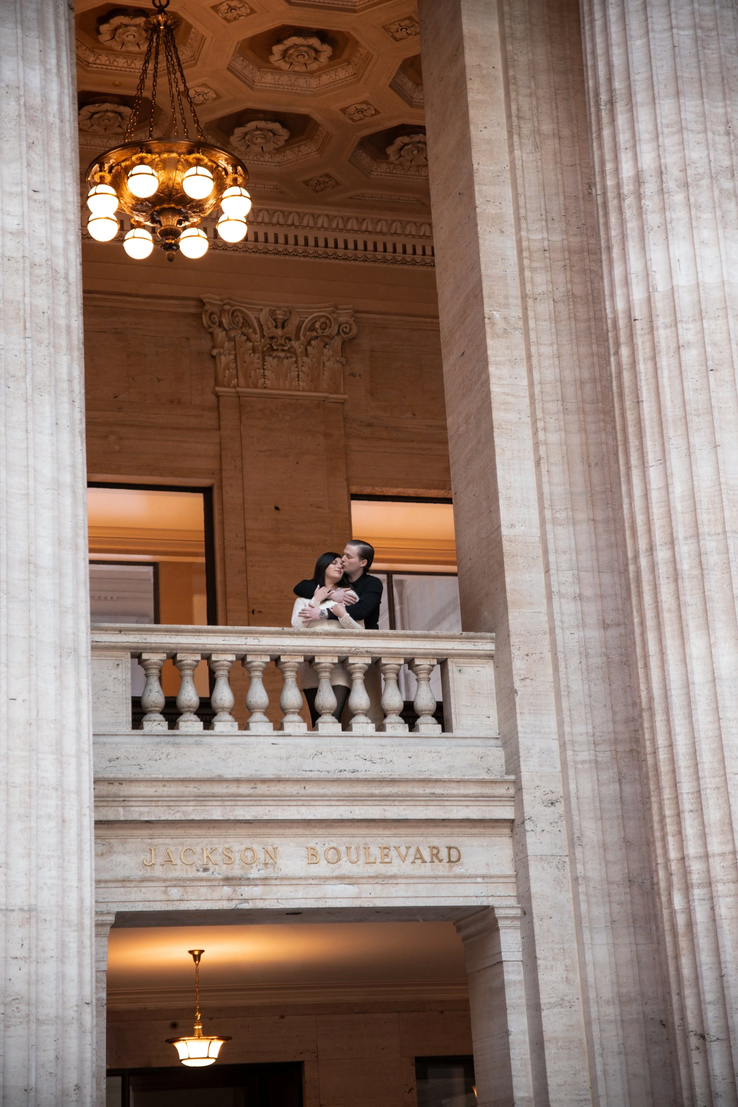 A couple kissing on a balcony inside a grand building with ornate pillars, chandeliers, and decorative ceiling features, with a sign that reads 'Jackson Boulevard'.