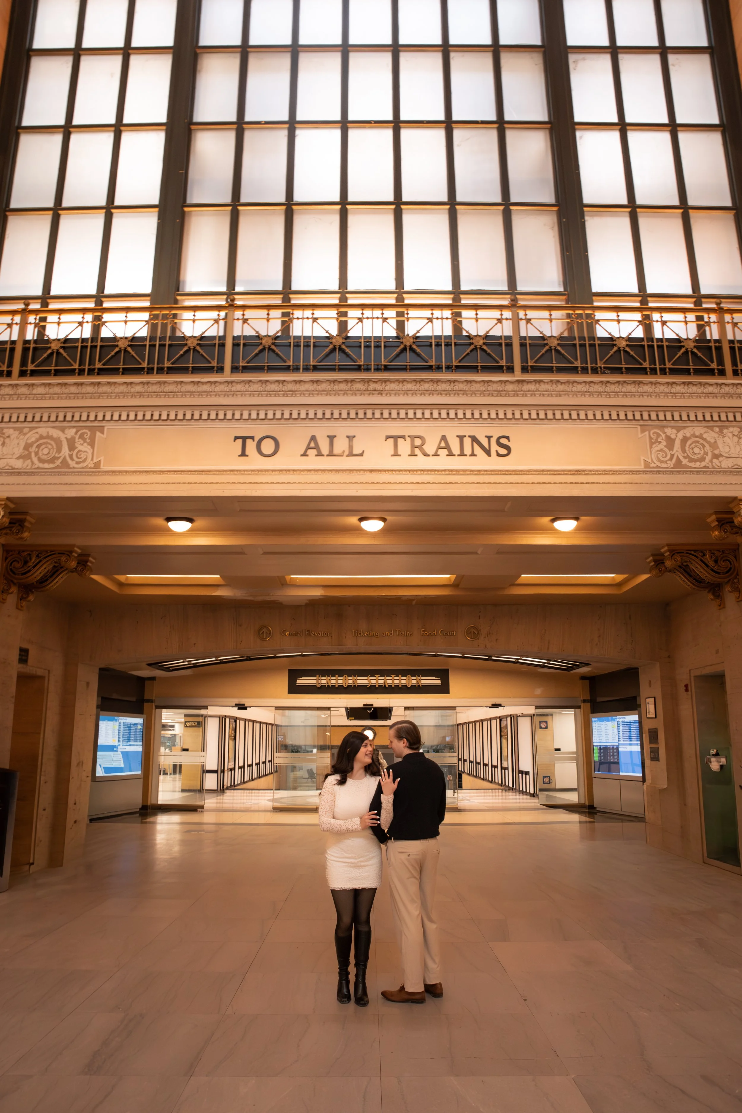 Interior of a train station with a sign reading 'To All Trains' and two people talking, standing in front of the ticketing and train information area.
