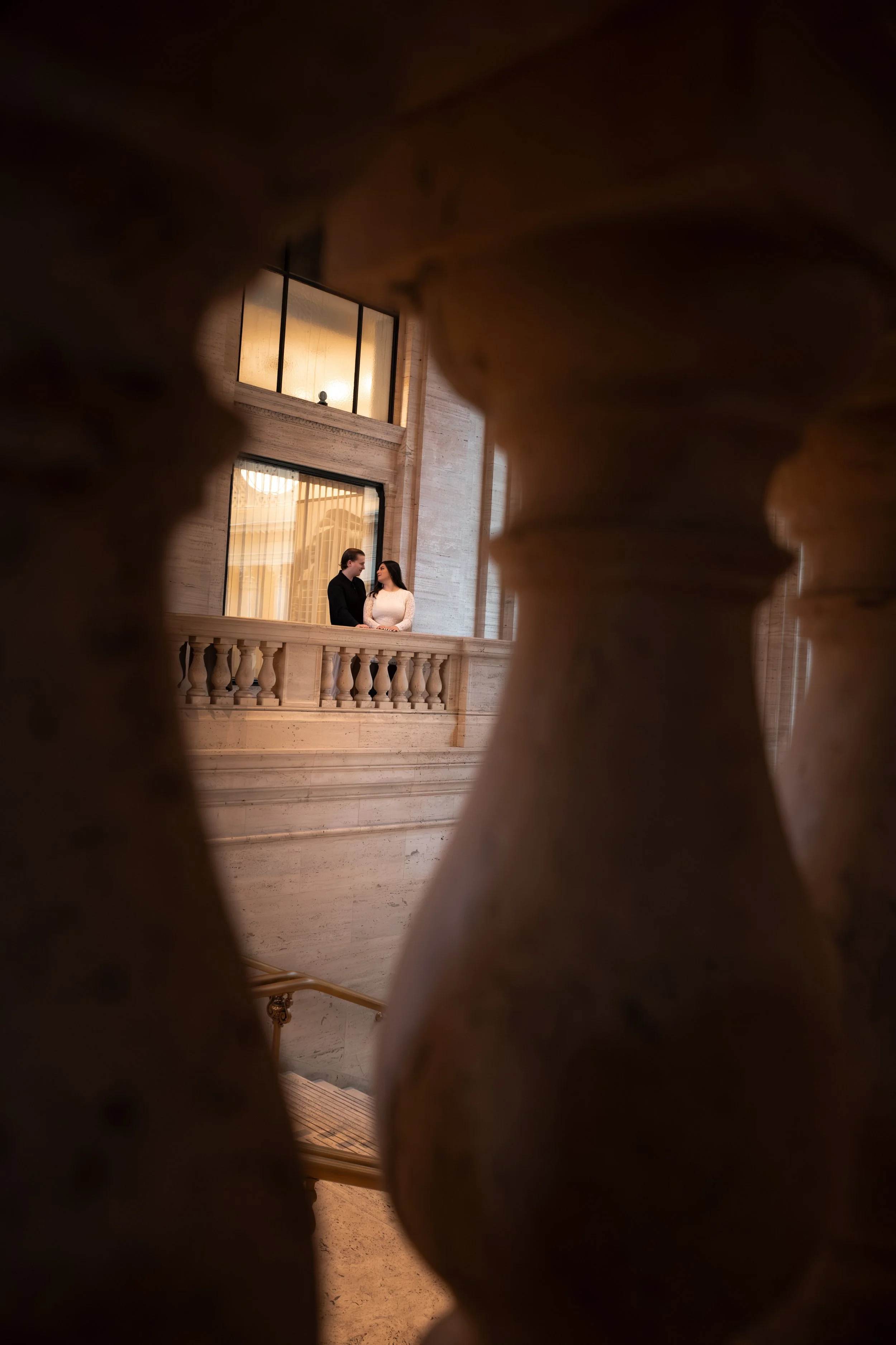 A couple standing together on a balcony, framed by a stone balustrade.