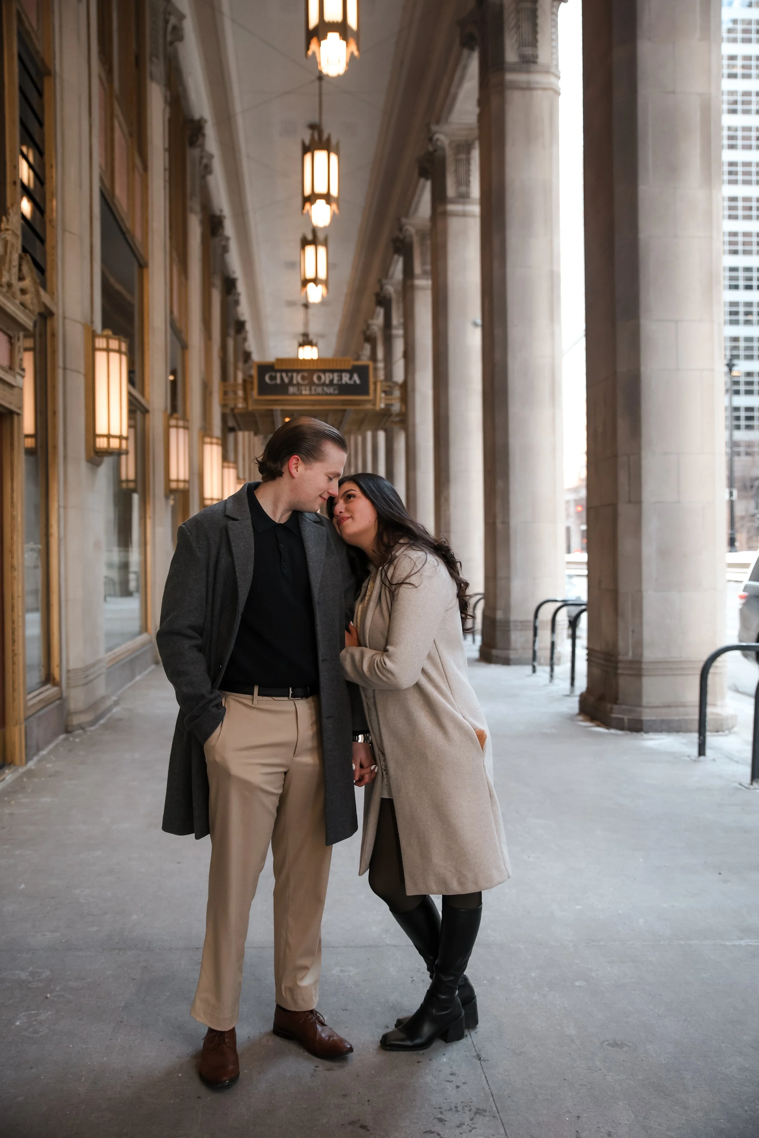 A man and woman standing close together under an arched, columned walkway outside a building labeled "Civic Opera." They are leaning their foreheads together, holding hands, and looking into each other's eyes, dressed in stylish winter clothing.