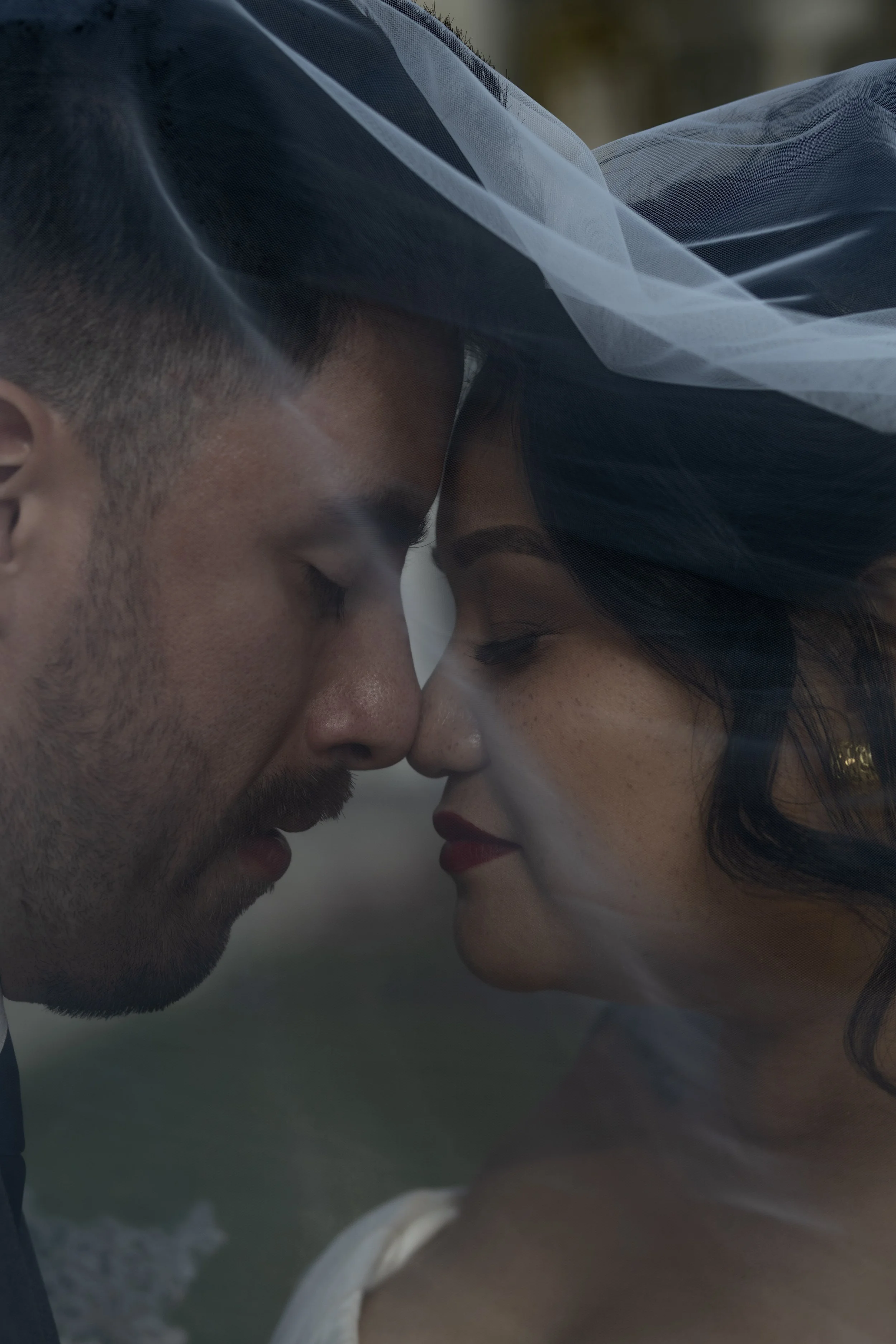A close-up of a man and a woman with their foreheads touching and eyes closed, wearing wedding attire with a veil and a white dress, outdoors near water.