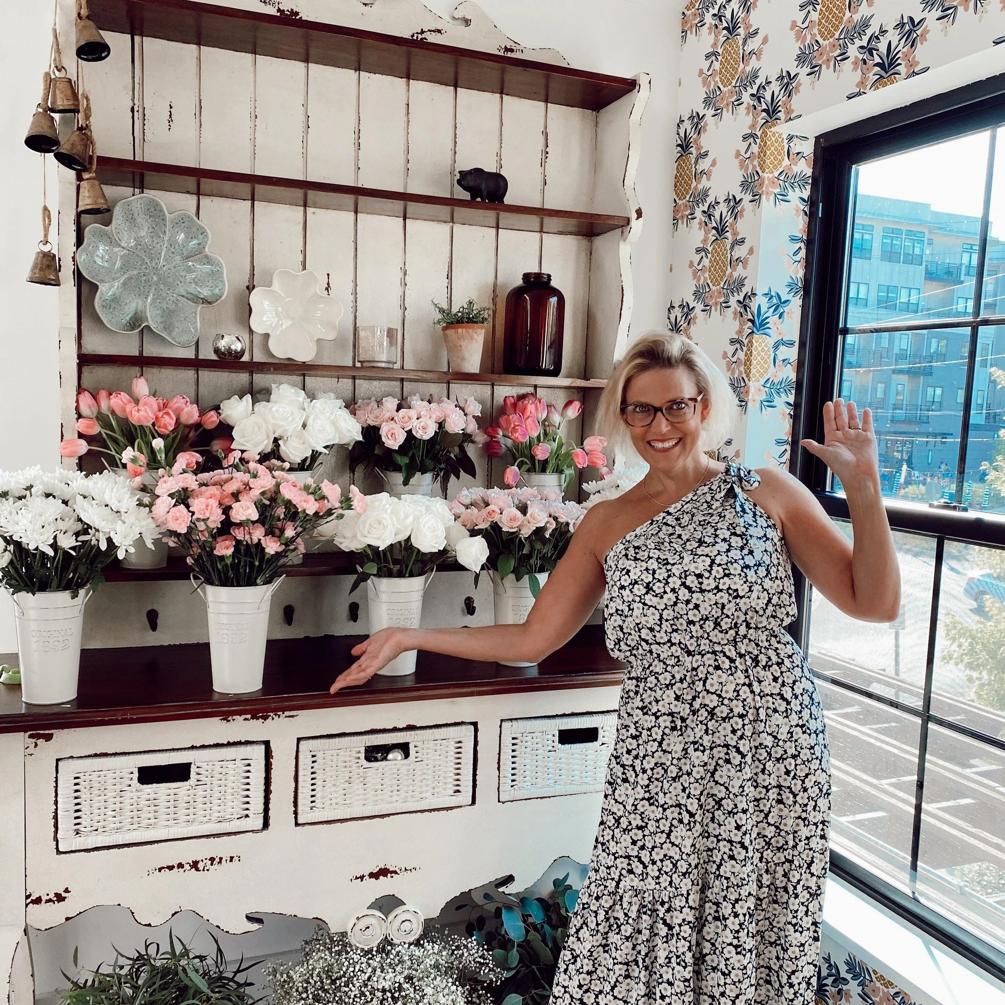A woman in a floral dress smiling and waving inside a floral shop, with pink and white flowers in white buckets on a distressed white shelf and large windows showing a city street outside.