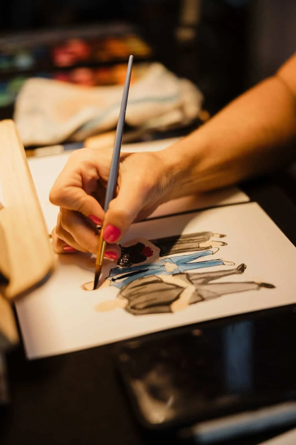A person painting wedding guests with watercolor, showing figures in blue, black, and beige tones.