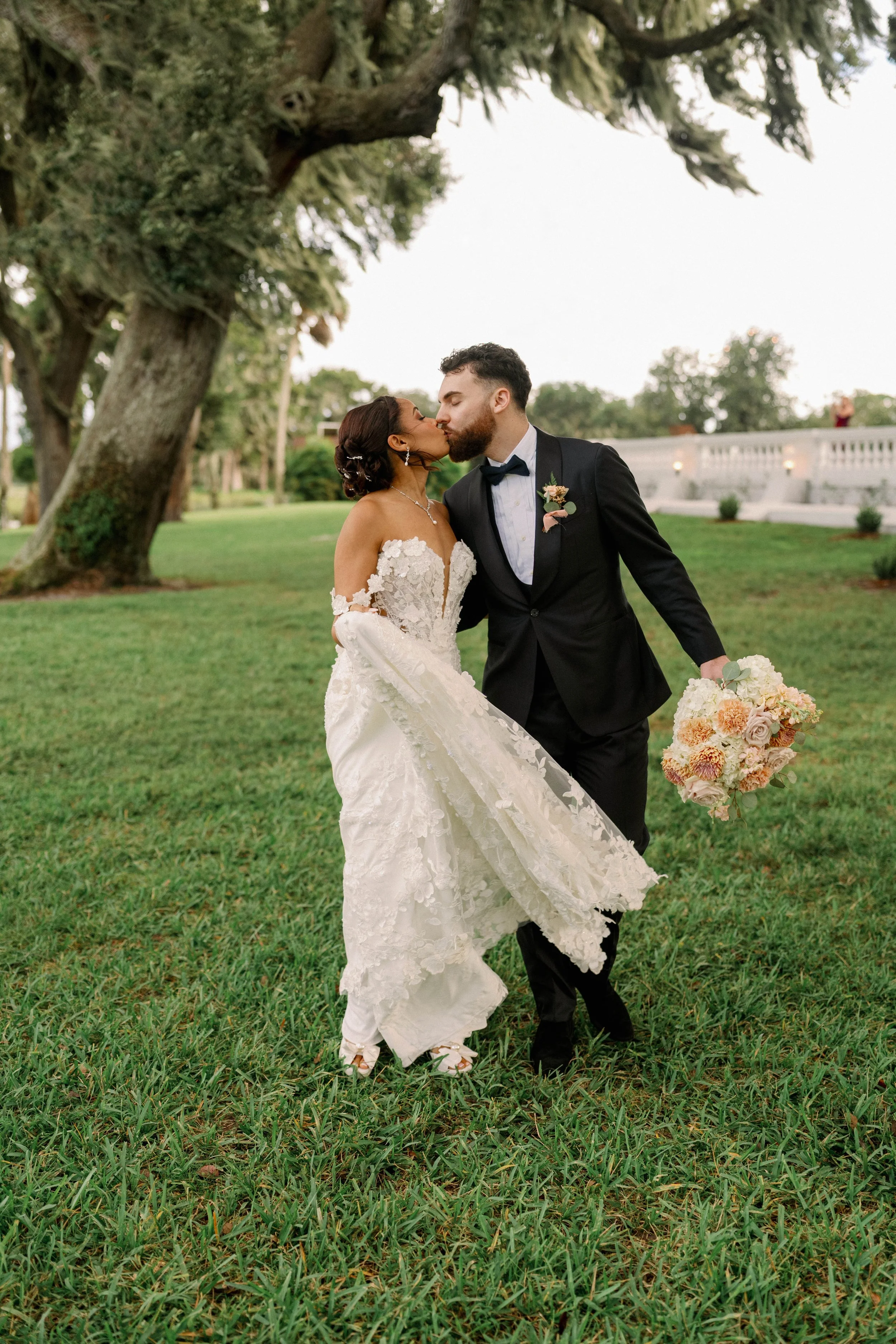 Lauren & Melvin share a kiss outdoors on their wedding day. The bride is in a white lace wedding gown holding a bouquet, and the groom is in a black tuxedo with a bow tie. They stand on green grass near a large tree.
