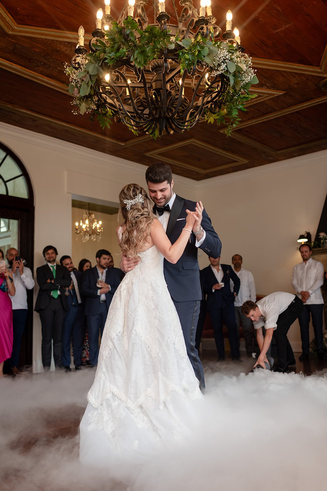 A couple having their first dance at a wedding under a decorative chandelier, surrounded by guests and smoke effects.