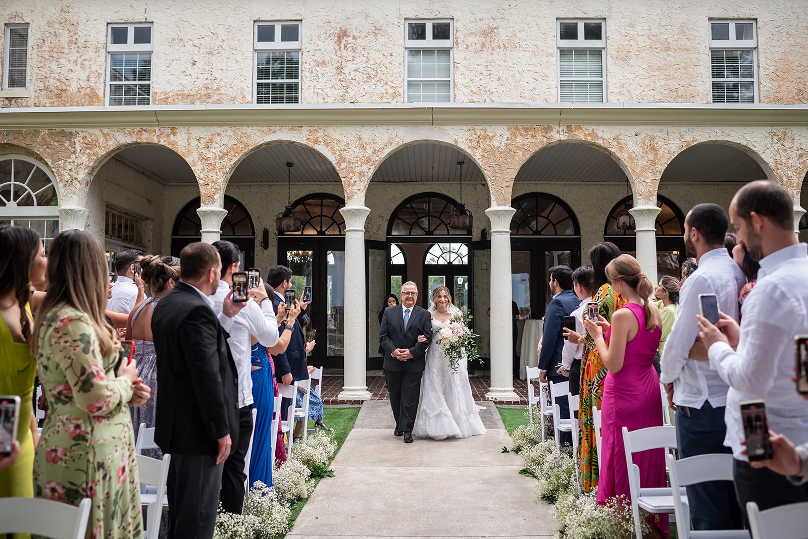 Bride walking down the aisle with her escort at an outdoor wedding ceremony, surrounded by guests taking photos with their phones.