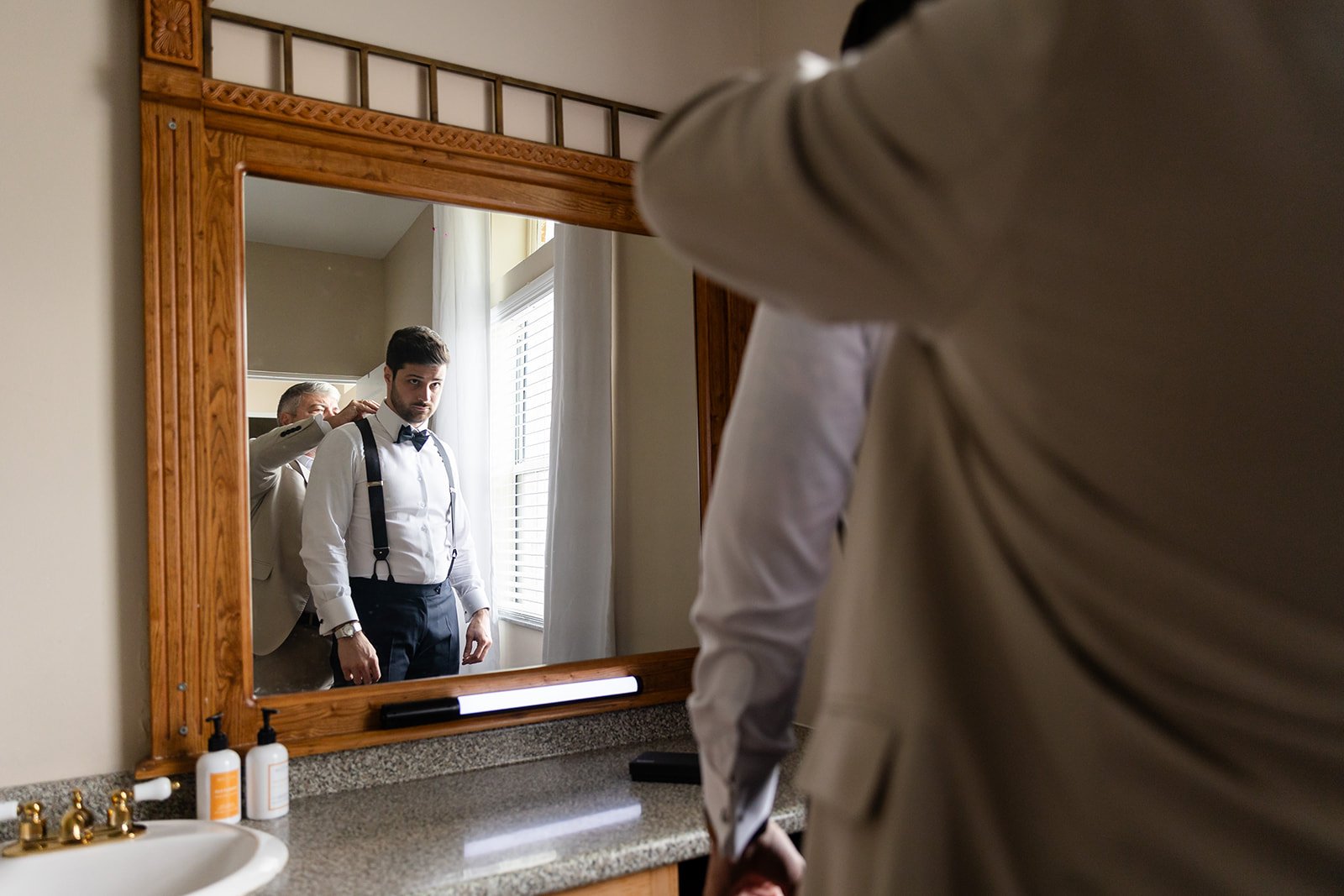 Two men getting dressed in formal attire in front of a large mirror. One is adjusting the other's suspenders.