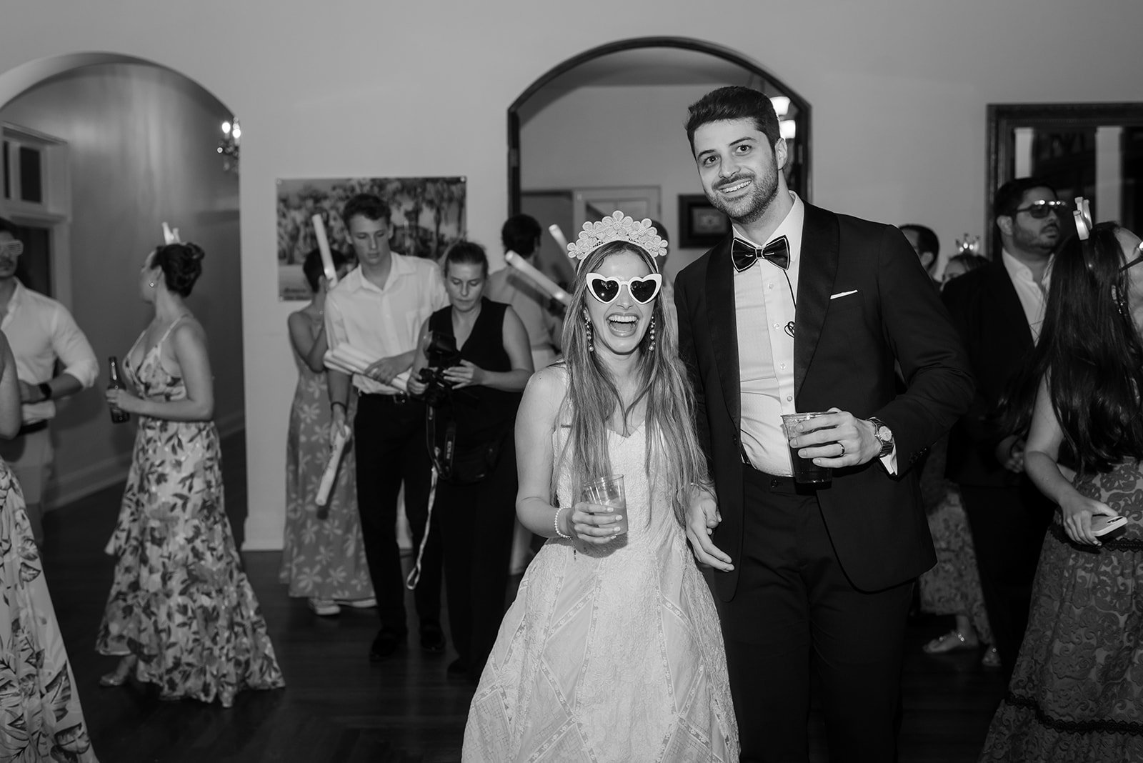 A black and white photo of a wedding reception with a bride in a dress and heart-shaped sunglasses next to a groom in a suit. Guests are socializing in the background, some holding drinks.