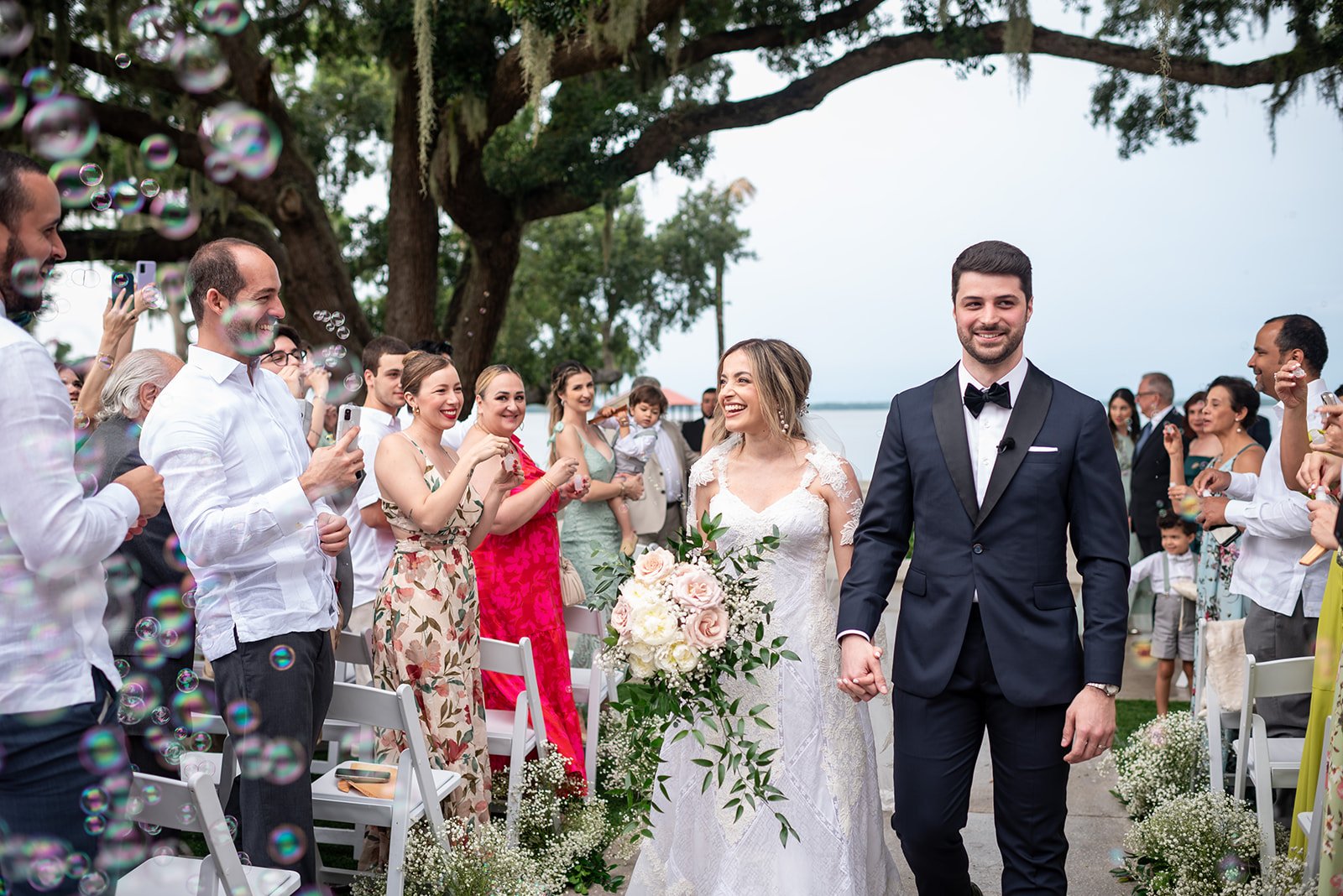 A bride and groom walking down the aisle outdoors, smiling, surrounded by joyful guests blowing bubbles.