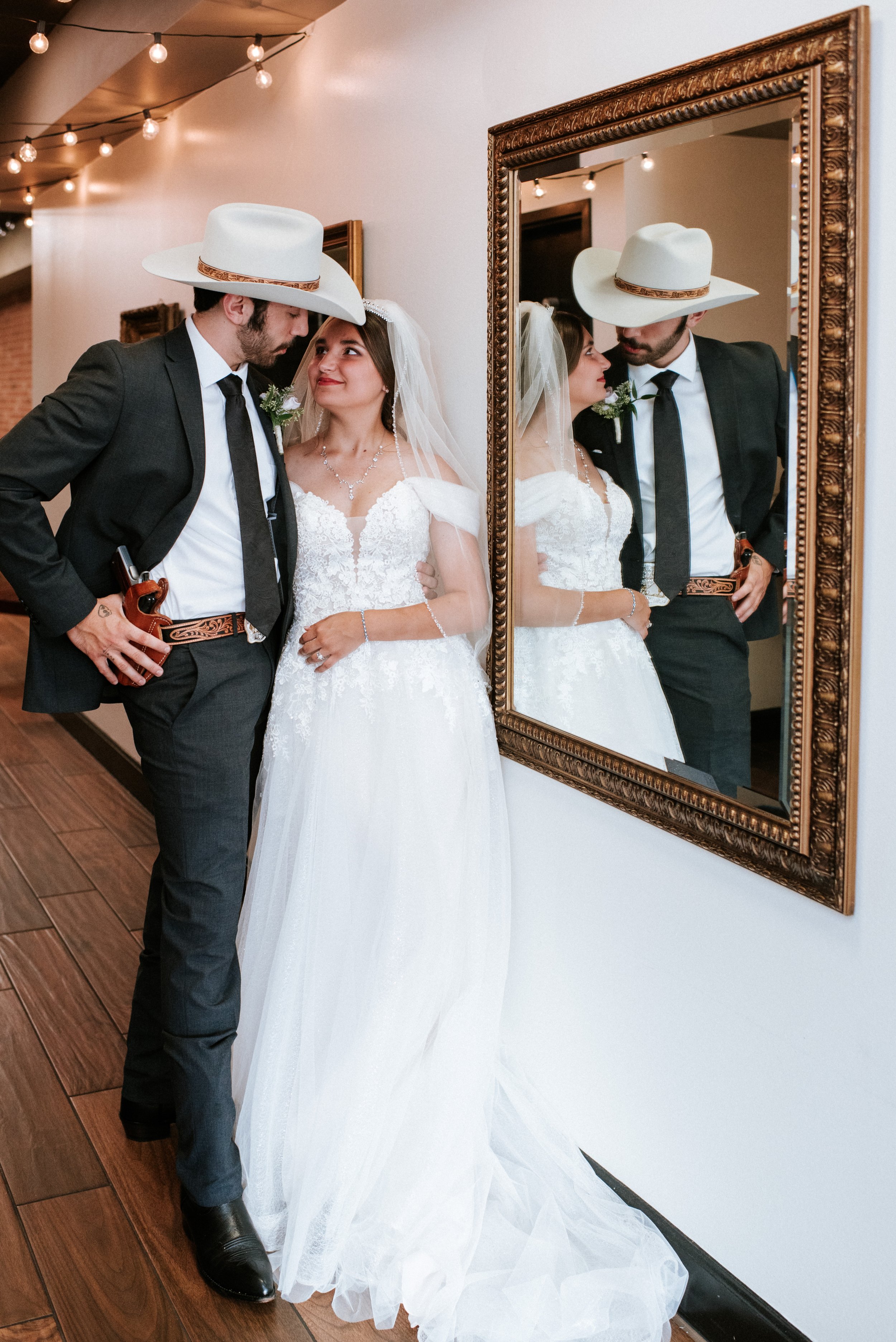 Bride and groom in formal attire with cowboy hats, standing by a mirror, decorated hallway, wooden floor, and string lights overhead.