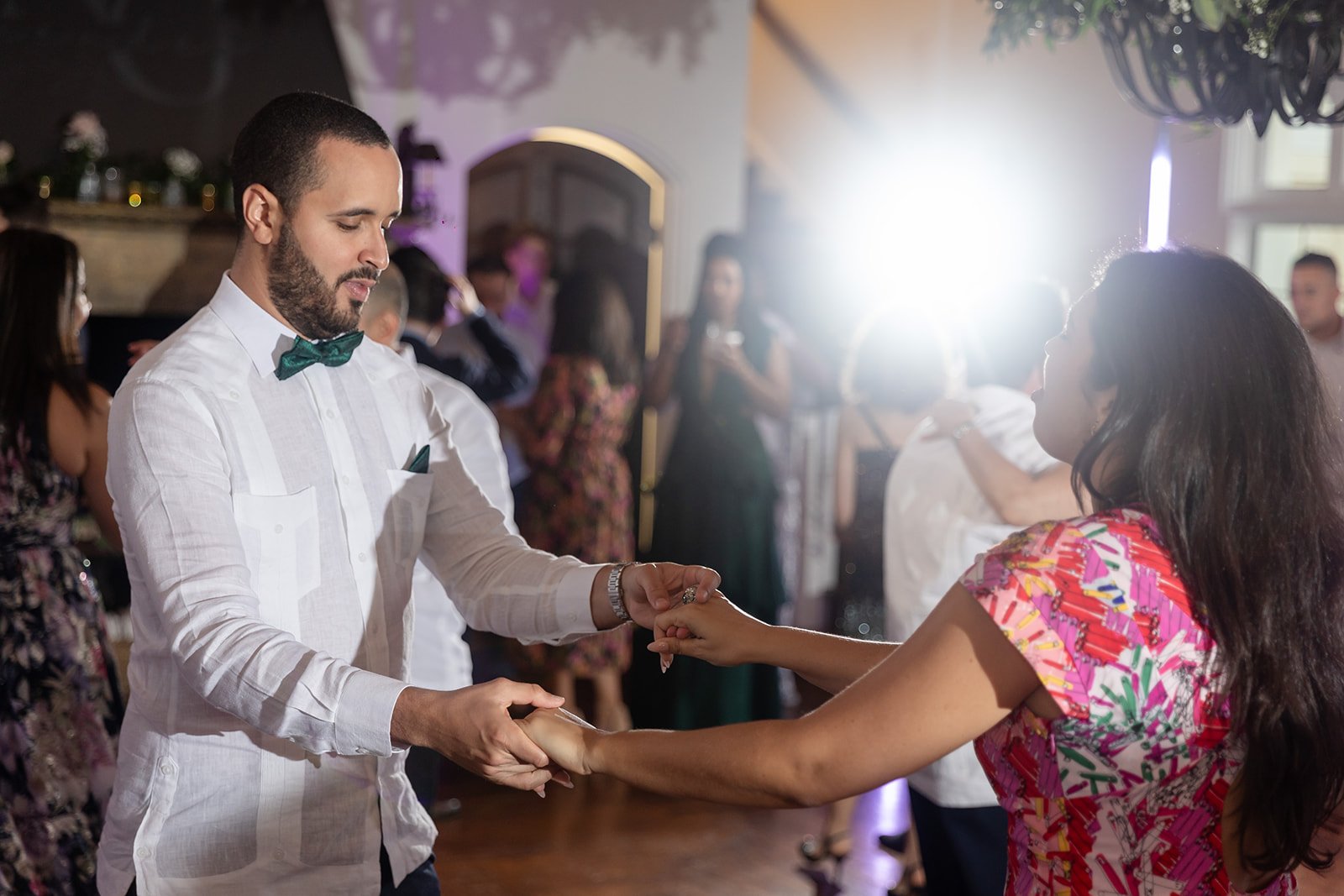 A man and woman dancing at an event, dressed in formal attire. The man is wearing a white shirt with a green bow tie, and the woman is wearing a colorful dress. Other people are also dancing in the background, with bright lighting illuminating the sc