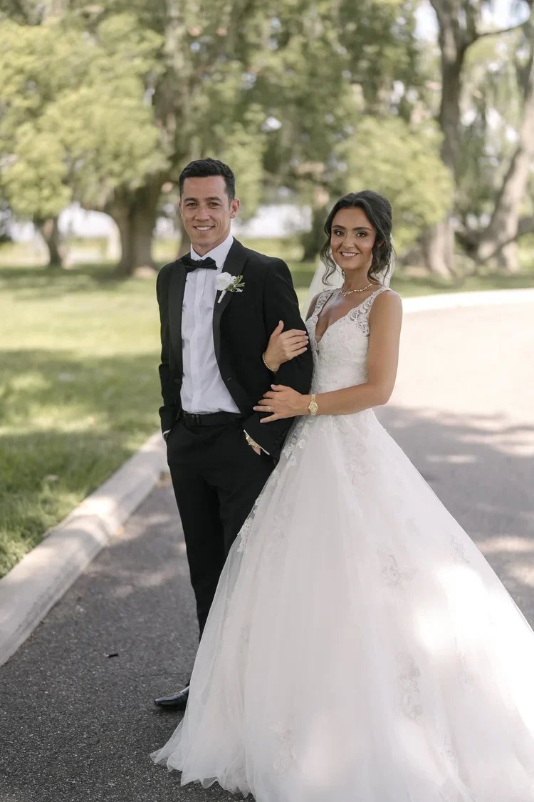 A bride and groom standing outdoors on a sunny day in a park with green trees in the background. The groom is wearing a black tuxedo with a bow tie and white shirt, and the bride is wearing a white wedding dress with lace details. The bride has her arm linked through the groom's arm, and both are smiling at the camera.