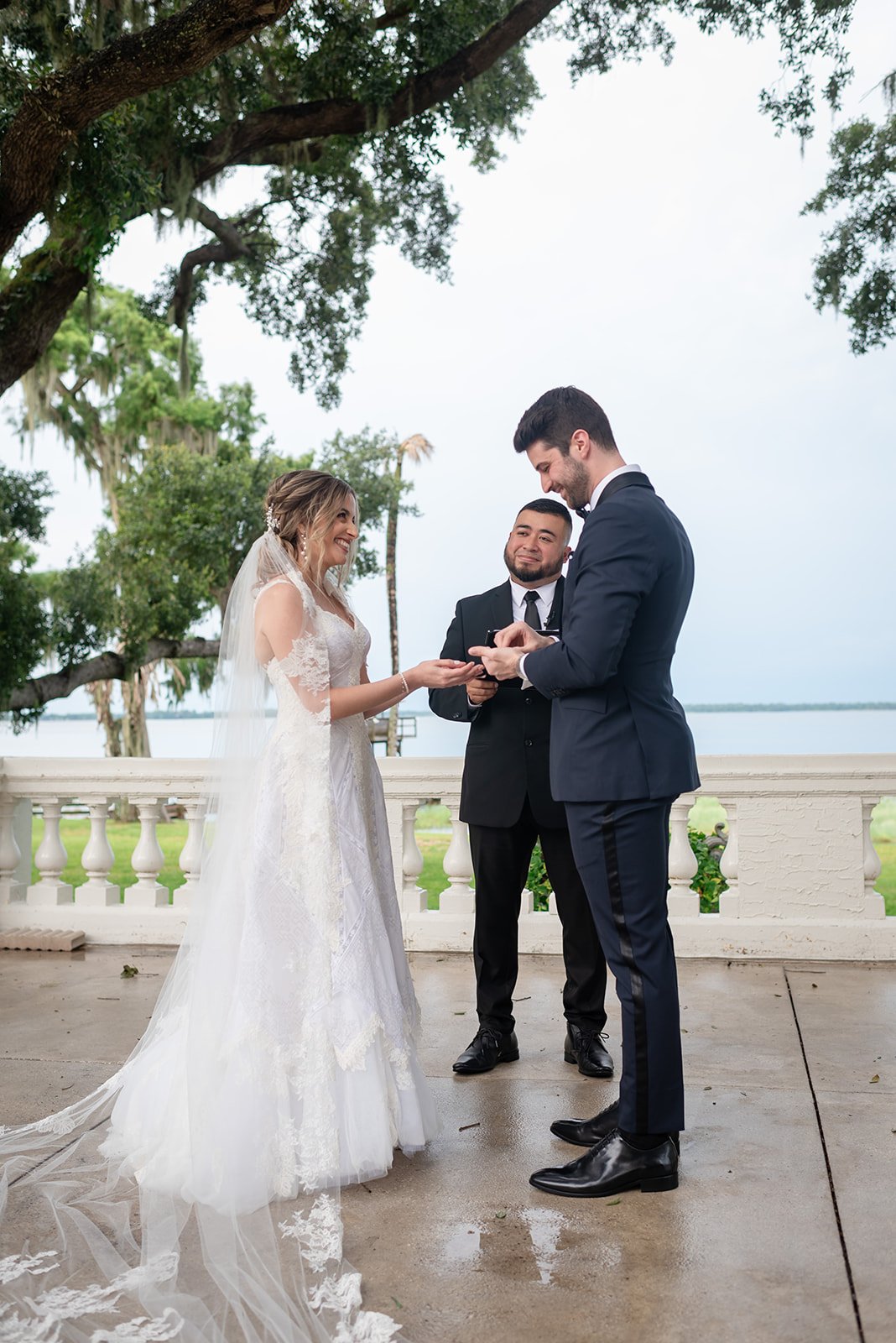 A bride and groom exchanging rings in an outdoor wedding ceremony with an officiant. The bride is wearing a lace wedding dress and veil, and the groom is in a suit. They are standing on a balcony with trees and water in the background.