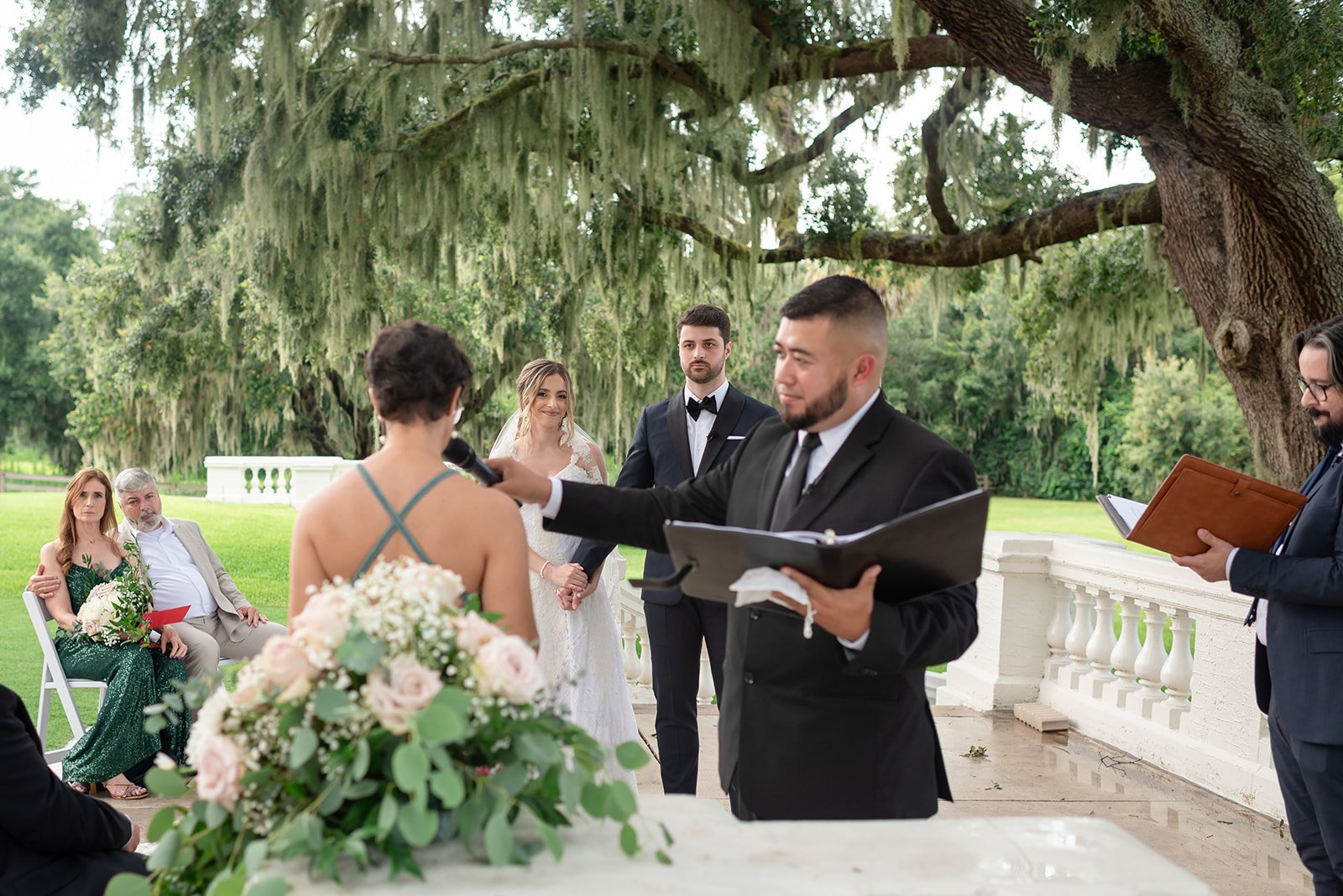 Outdoor wedding ceremony with an officiant speaking, a bride in a white dress, a groom in a tuxedo, and guests seated under a tree with hanging moss.