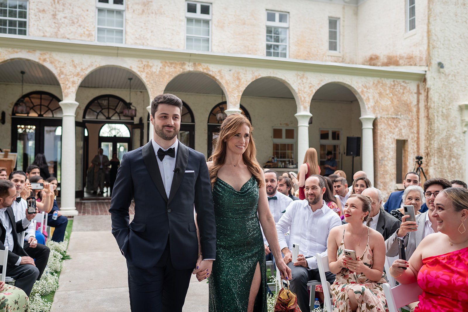 A man in a tuxedo and a woman in a green sequined dress walk down an aisle at an outdoor event, possibly a wedding ceremony. They are holding hands and smiling, surrounded by seated guests who are watching and taking photos. The venue features an ele