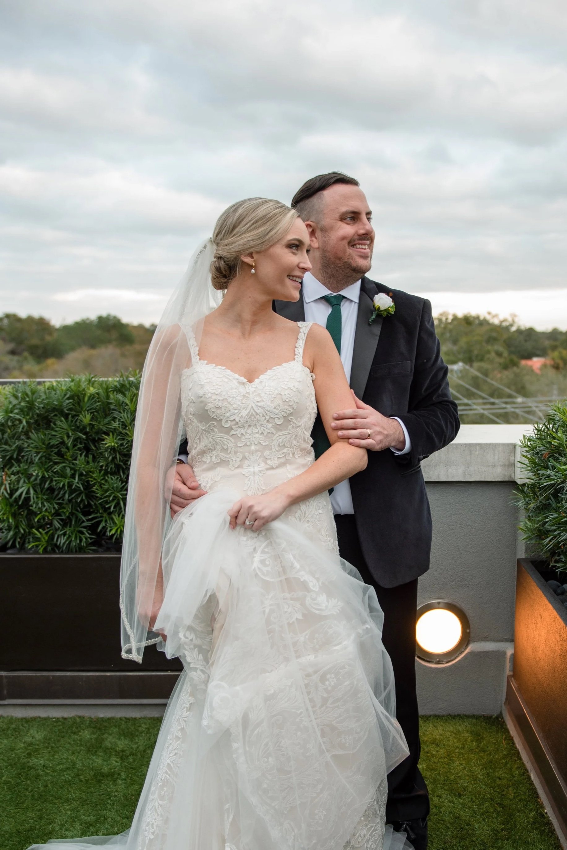 A bride and groom stand on an outdoor terrace, smiling and embracing, with cloudy skies and trees in the background. The bride wears a lace wedding dress with a veil, and the groom wears a dark suit with a white shirt and teal tie.