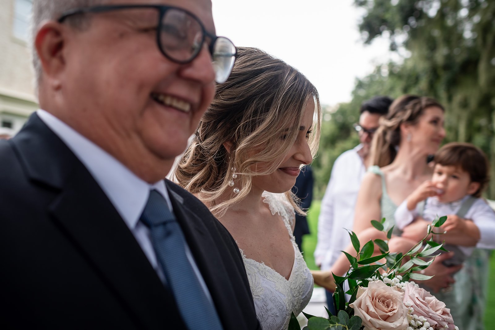 Bride walking with an older gentleman at an outdoor event, holding a bouquet of flowers, with other people in the background.
