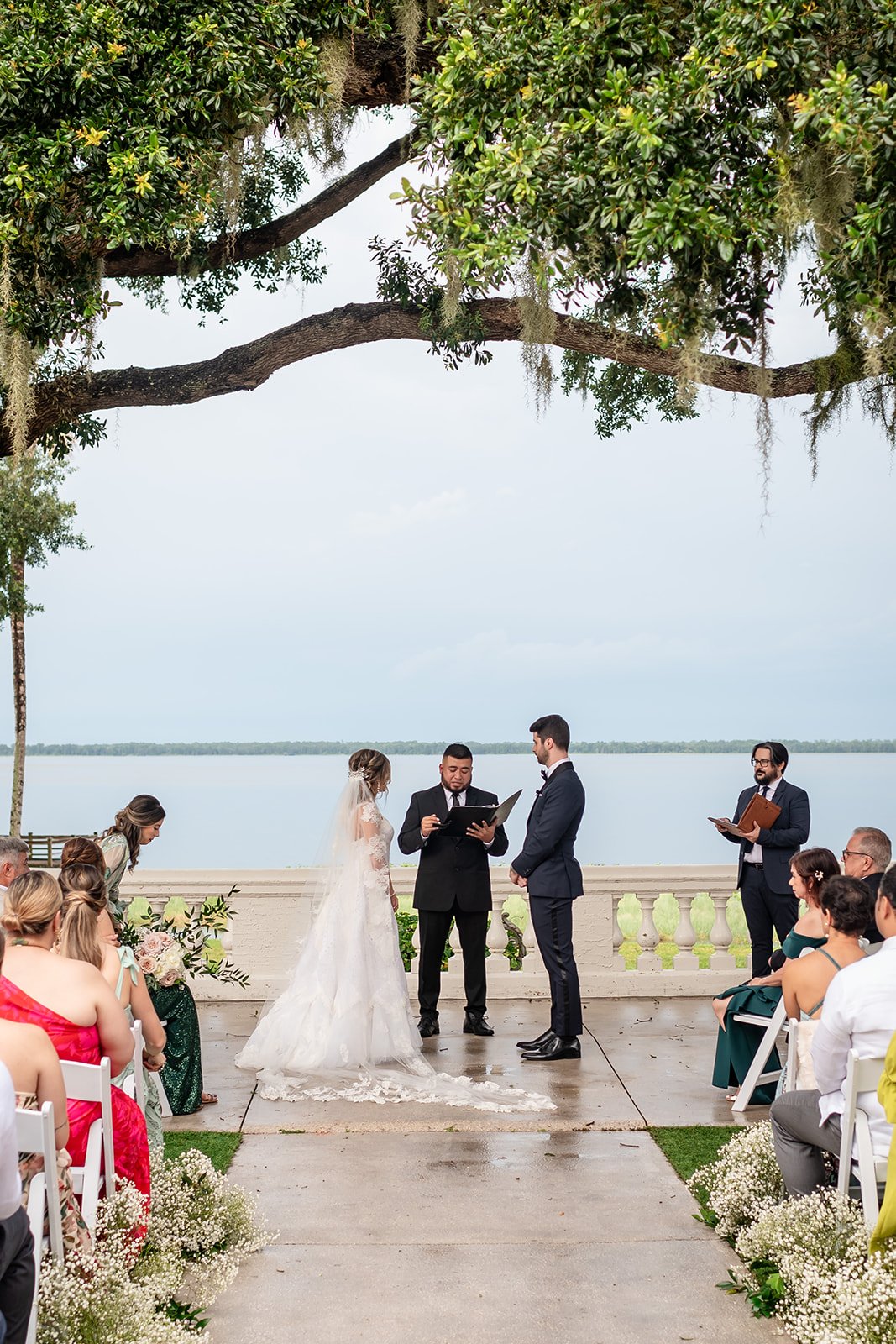 Outdoor wedding ceremony by a lake with bride, groom, officiant, and guests seated. Large tree branches overhead and floral decorations along the aisle.