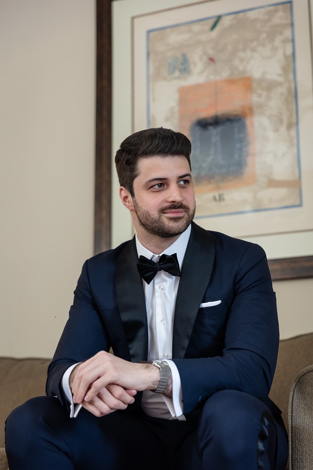 Man in a tuxedo sitting with hands clasped, looking to the side, with abstract art in the background.