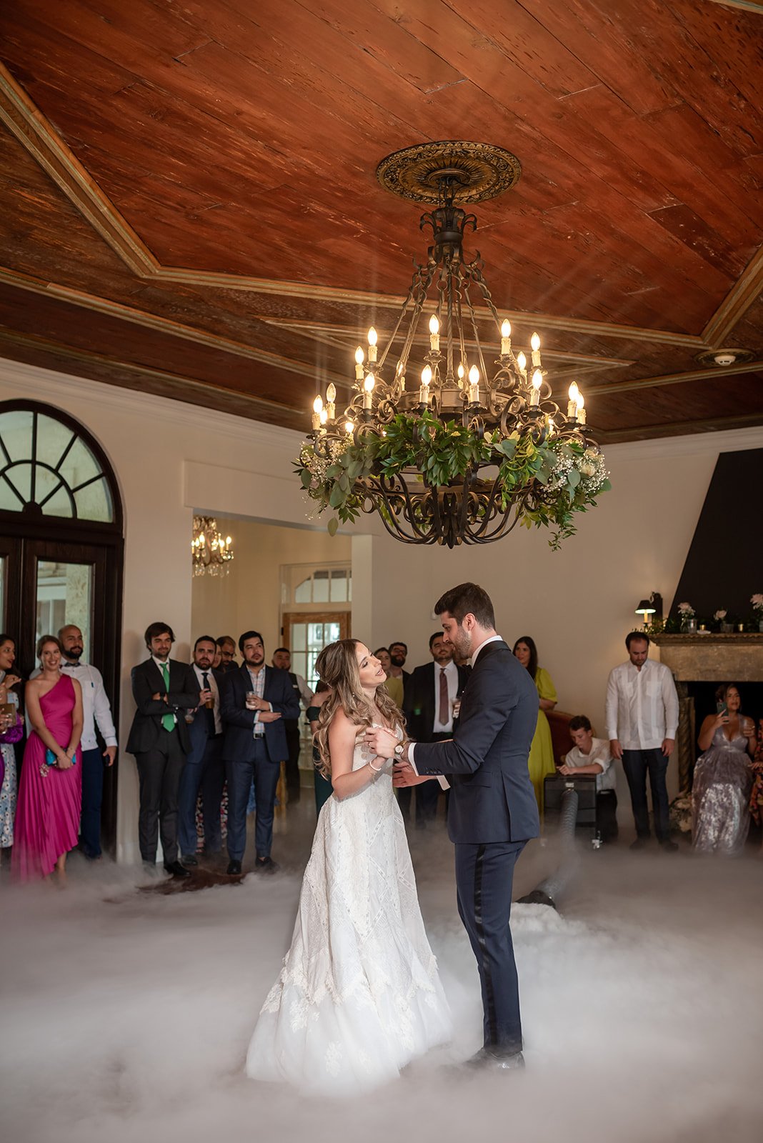 Bride and groom dancing under a chandelier with guests watching in an elegant venue.