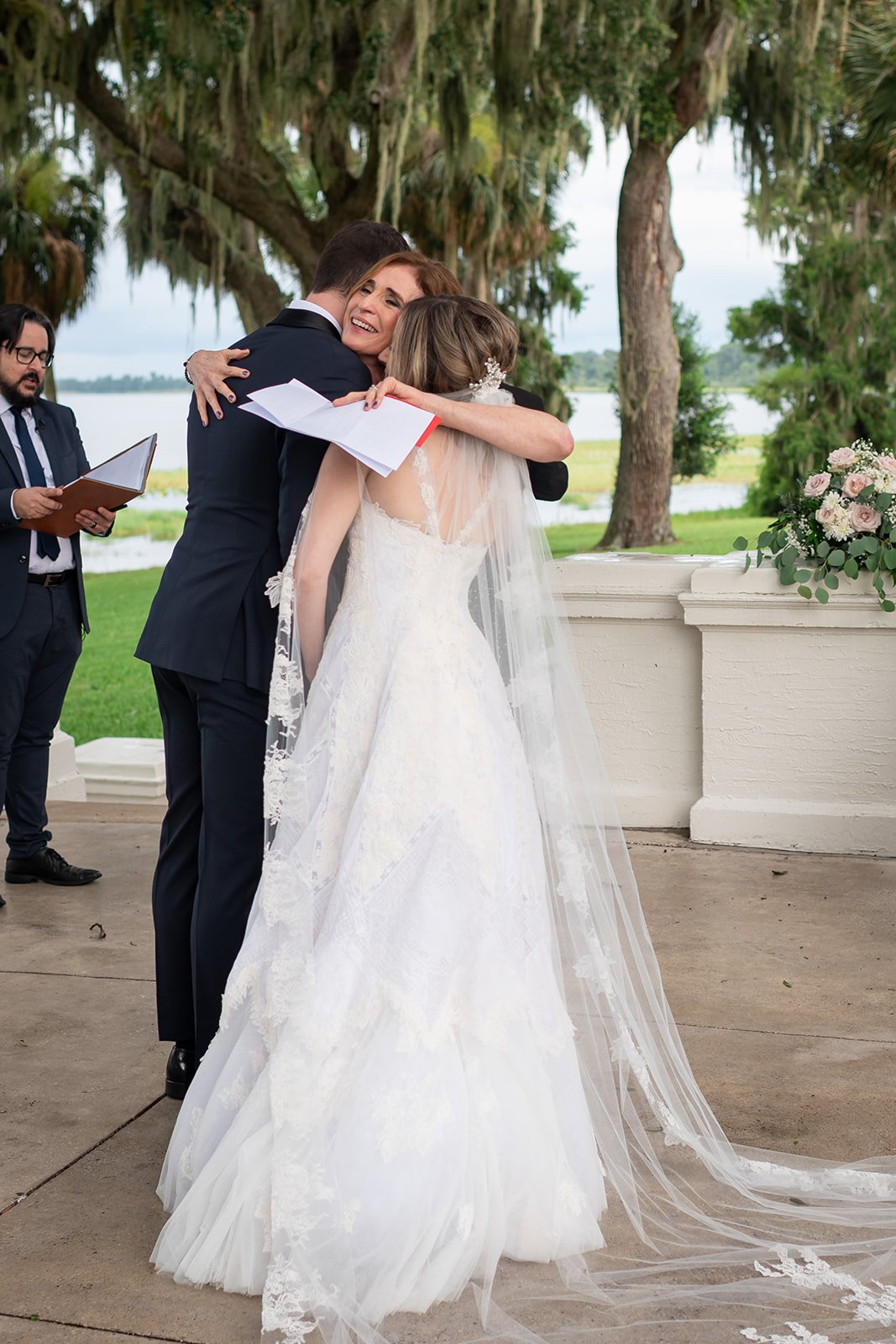 Bride and groom hugging a woman at an outdoor wedding ceremony with an officiant nearby.