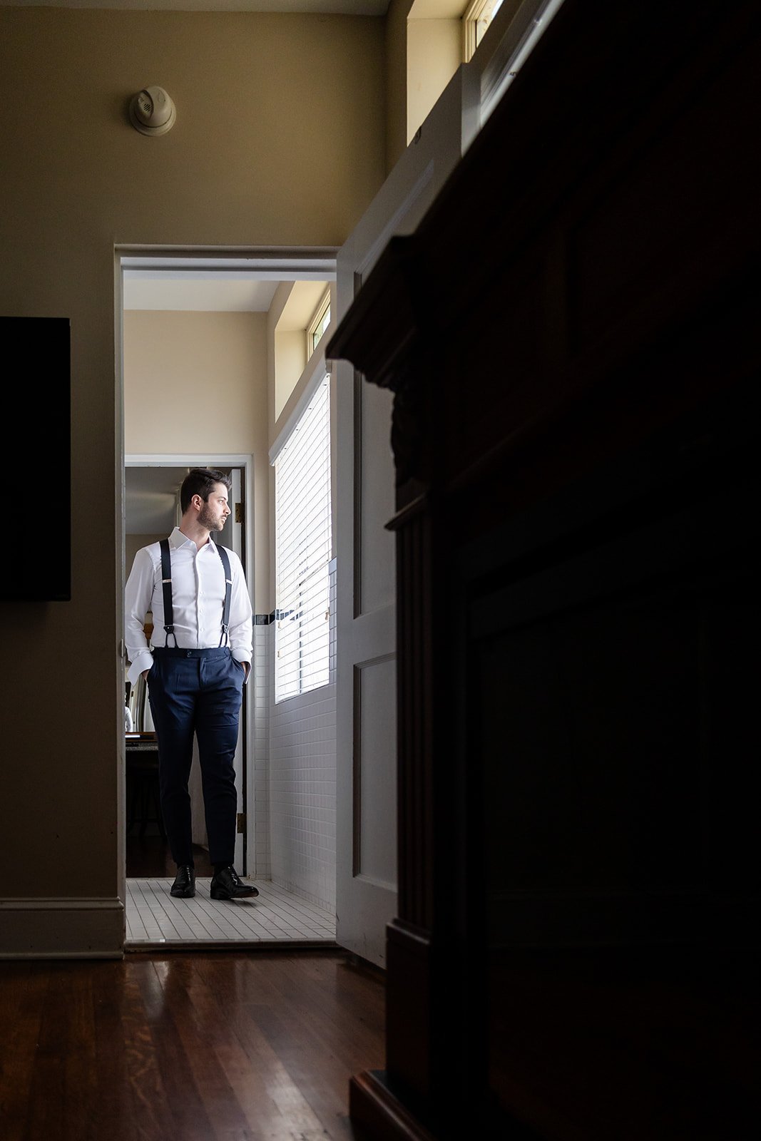 Man in formal attire standing in doorway, looking outside, in interior setting with wooden floor and wall vents.