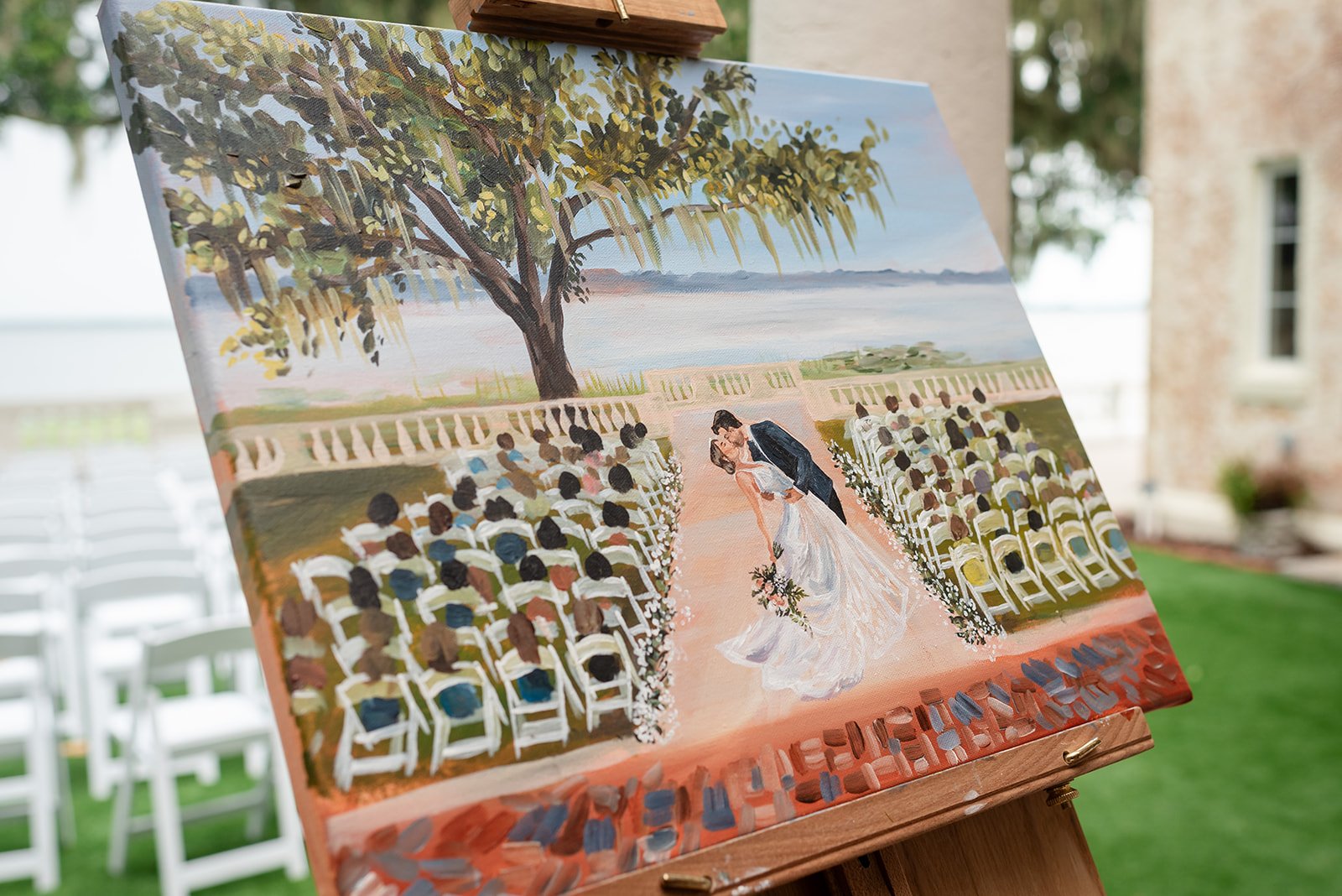 Painting on an easel depicting a wedding ceremony with a bride and groom kissing under a tree with guests seated on white chairs.