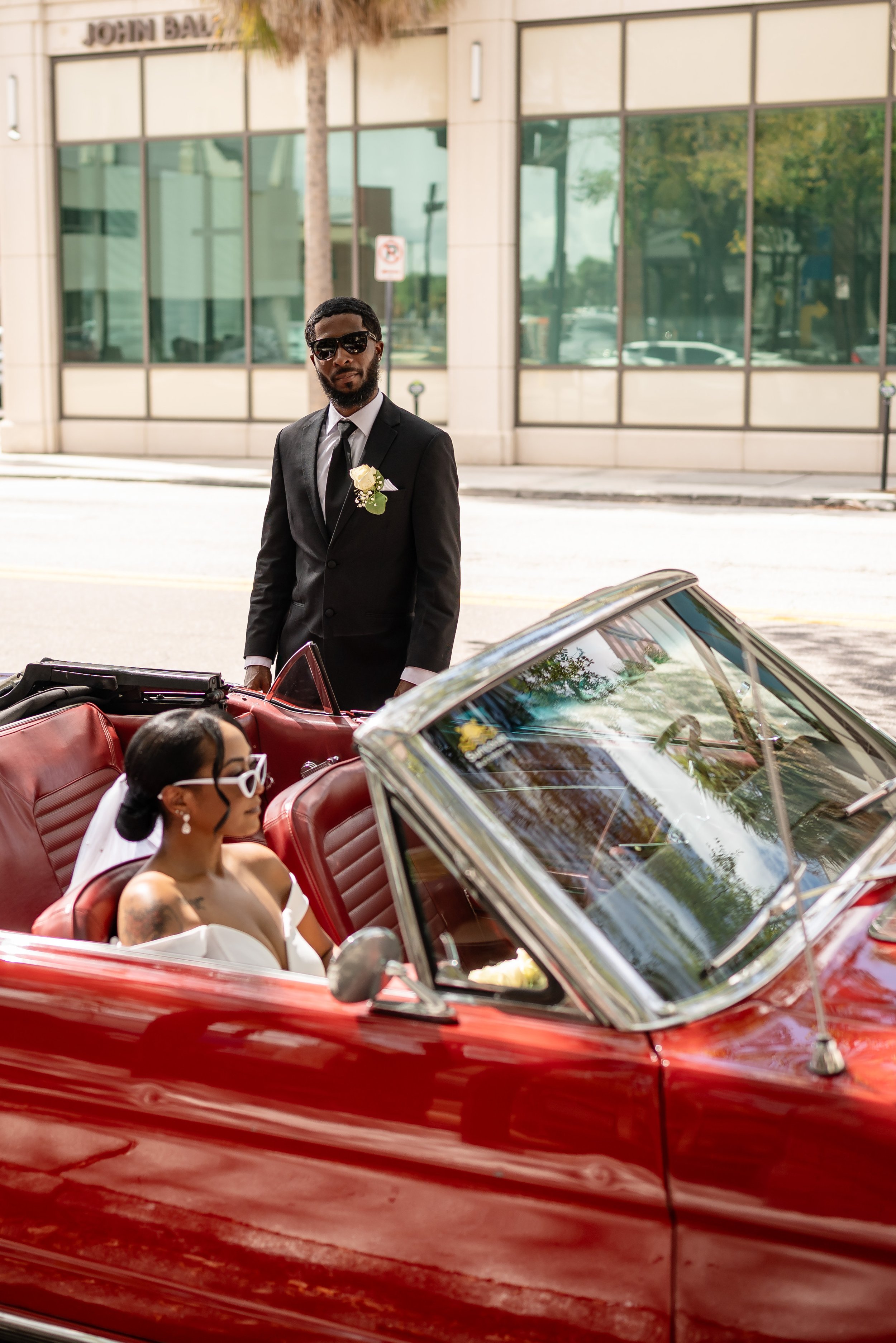Man in a suit with sunglasses and a boutonniere stands next to a woman in a red convertible wearing sunglasses and a wedding dress. Urban background with building reflections.