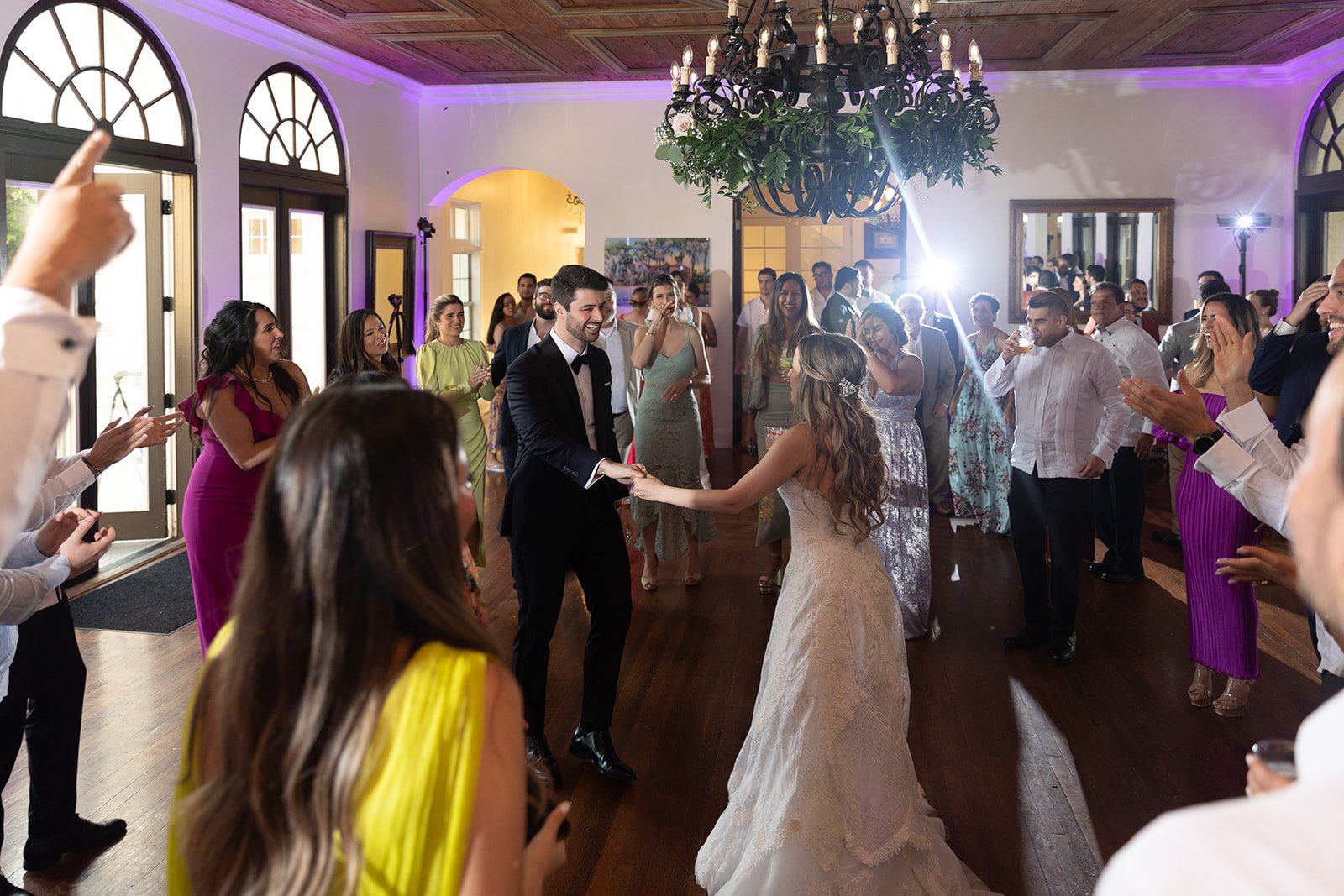 A couple dancing at a wedding reception surrounded by guests. The bride wears a white gown, and the groom is in a black suit. Guests are clapping and cheering in a decorated venue with a chandelier overhead and large windows.