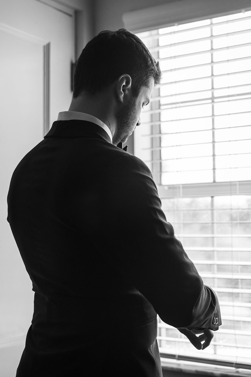 Black and white image of a man in a suit adjusting his cuffs, standing by a window with blinds.