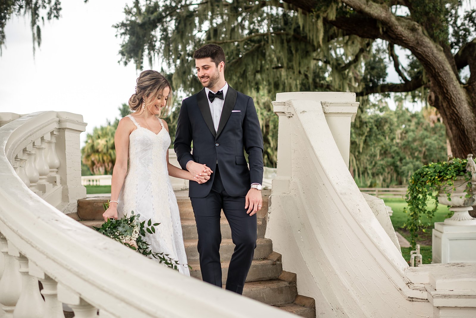 Bride and groom walking down stairs holding hands, surrounded by greenery and white stone banister, outdoor setting.
