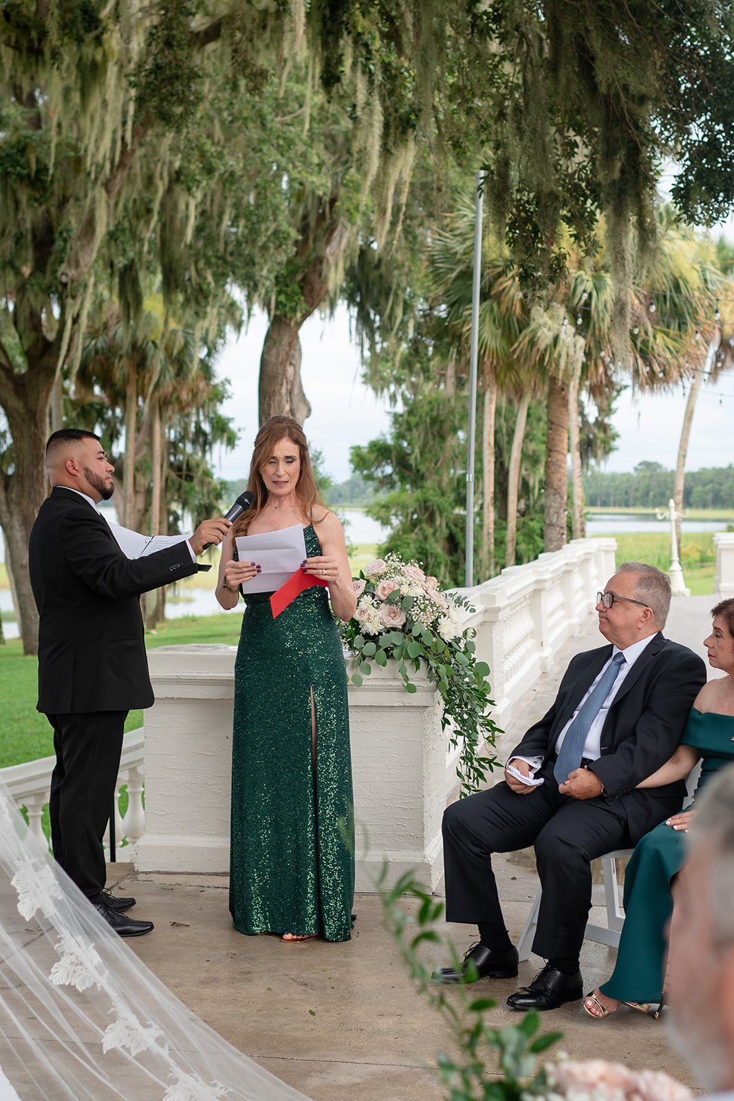 A woman in a green dress reads from a paper outdoors, with a man holding a microphone. Two seated individuals, a man and woman, listen attentively. Decorative flowers and trees in the background.