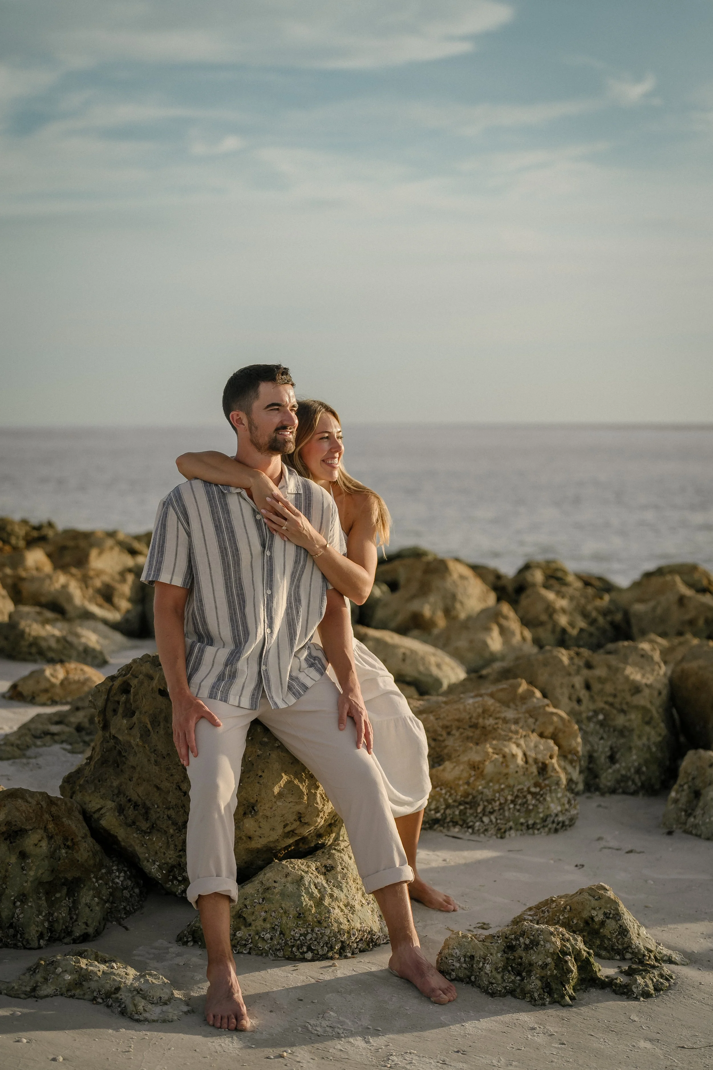 A happy couple sitting on rocks at the beach during sunset, smiling and enjoying the view.