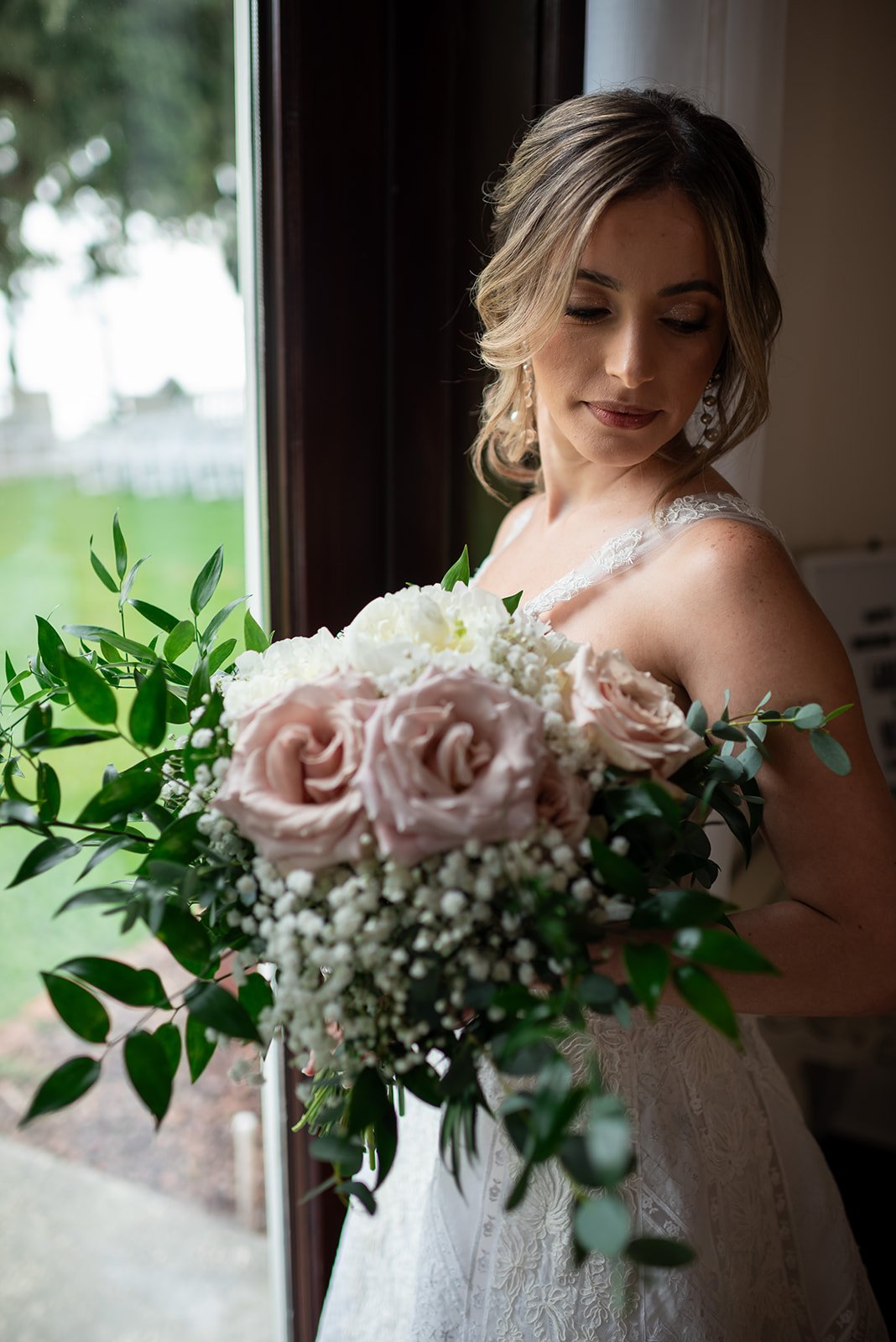 Bride holding a bouquet of pink roses and white flowers by a window.